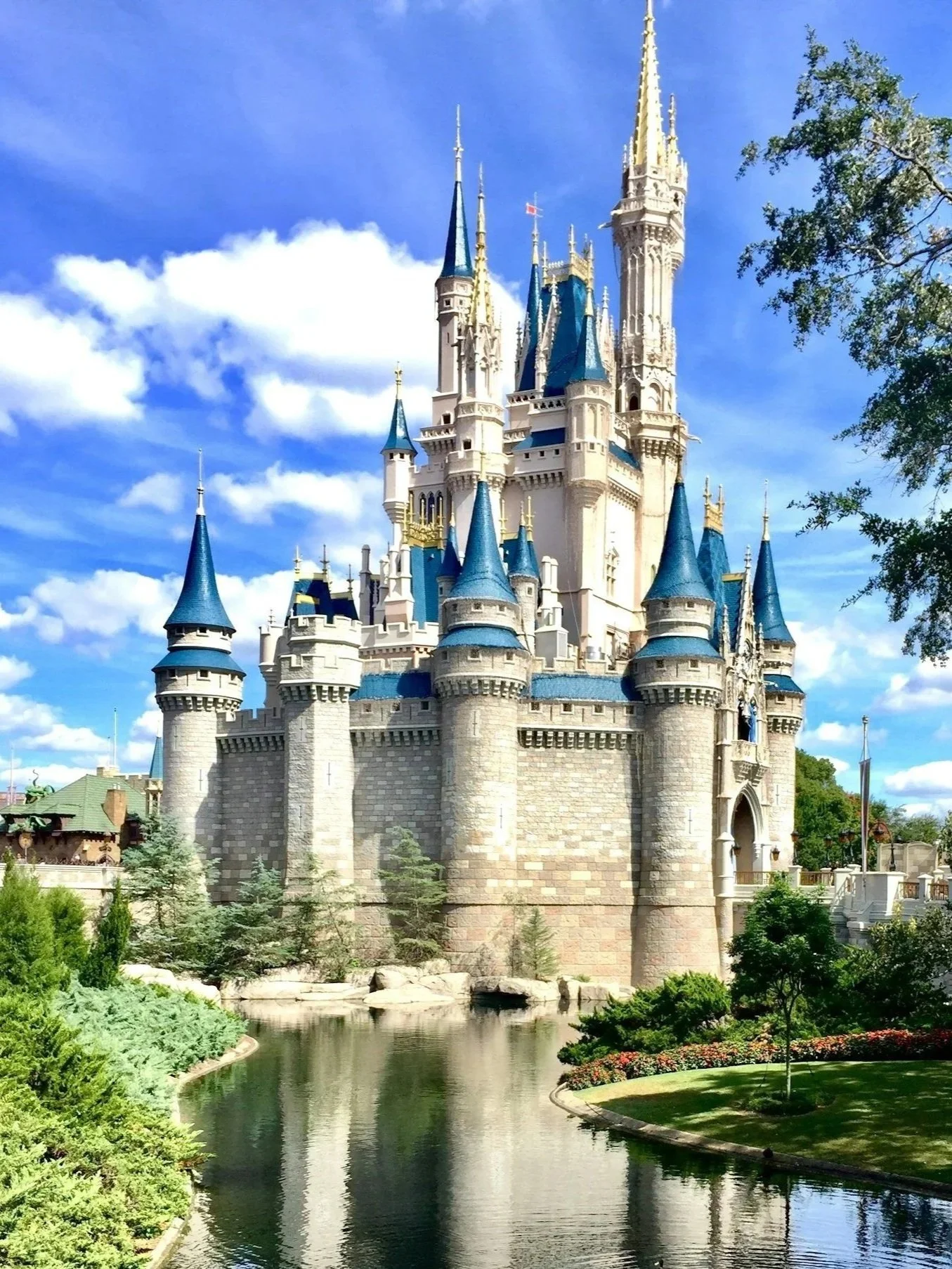 A large fairy tale castle with blue roofs and tall spires, surrounded by a moat and lush greenery, under a partly cloudy sky.