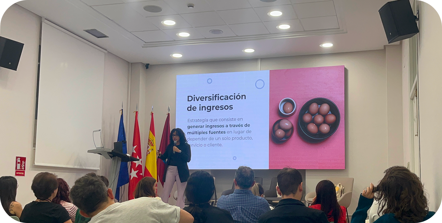 Persona ofreciendo una charla en un auditorio, con una diapositiva proyectada que dice “Diversificación de ingresos” junto a una imagen de varios huevos en un recipiente. El público está sentado observando.