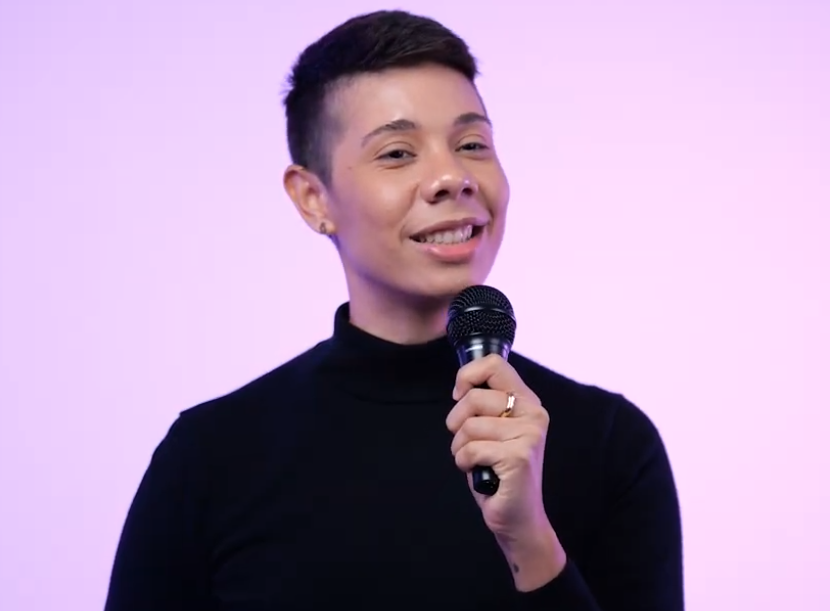 Layshi Curbelo con cabello corto sosteniendo un micrófono y sonriendo mientras habla, frente a un fondo lila suave. Lleva un suéter negro de cuello alto.