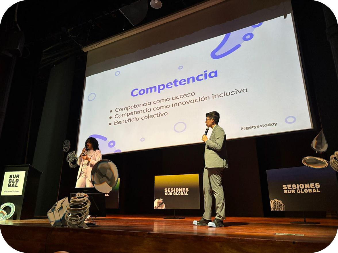 Dos personas de pie en un escenario dando una presentación frente a una pantalla grande con la palabra “Competencia” y una lista de puntos. El lugar tiene iluminación teatral y letreros de “Sesiones Sur Global”.