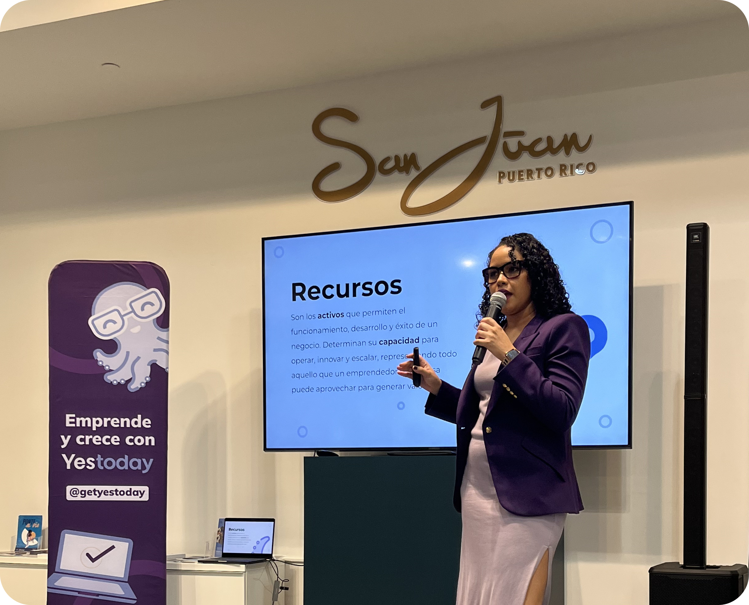 Persona hablando con micrófono frente a una pantalla que muestra la diapositiva “Recursos”. A la izquierda hay un banner morado de Yestoday y en la pared se lee “San Juan Puerto Rico”.