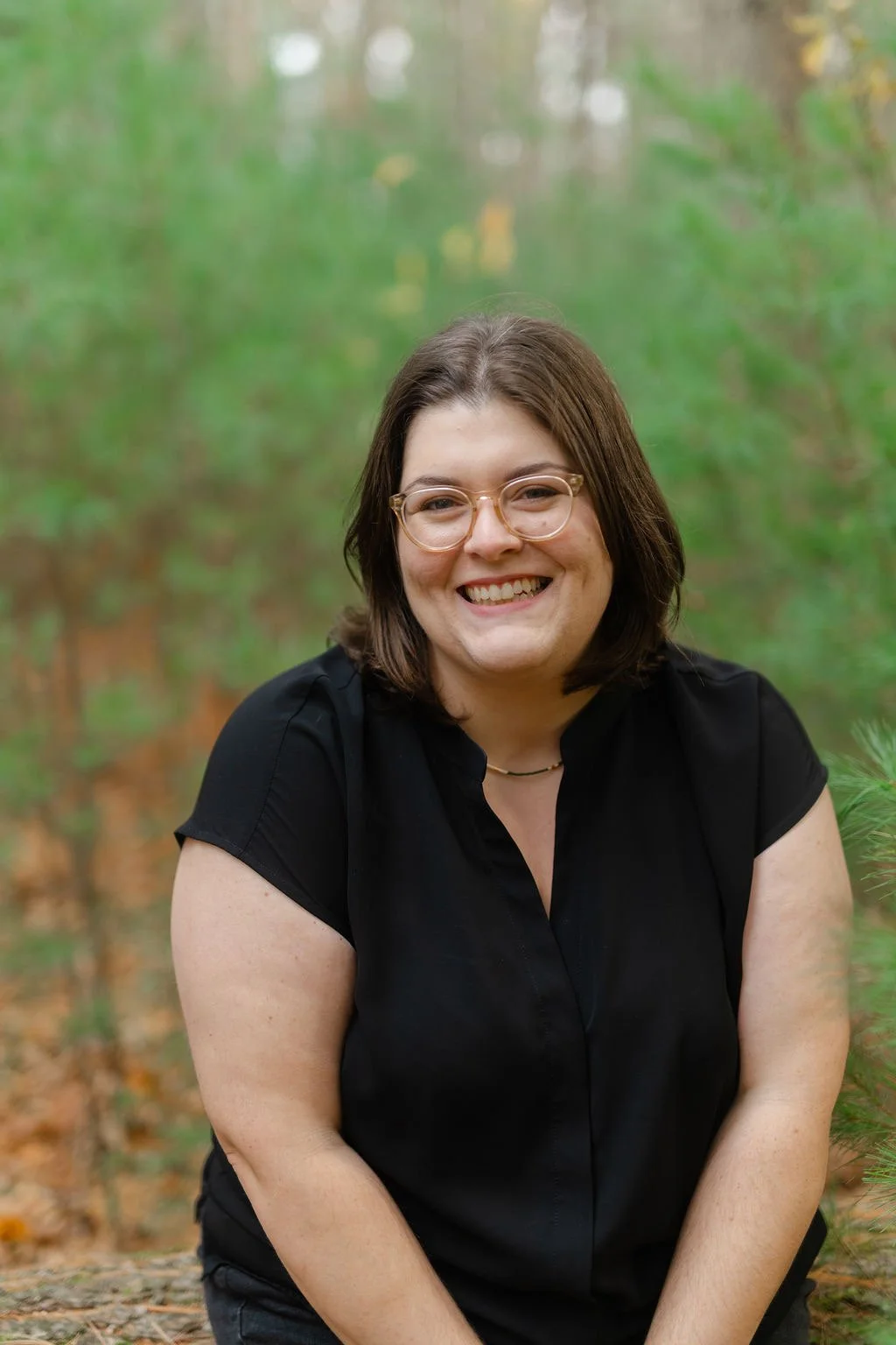 A woman with shoulder-length brown hair, glasses, and a black shirt smiling outdoors with green trees in the background.