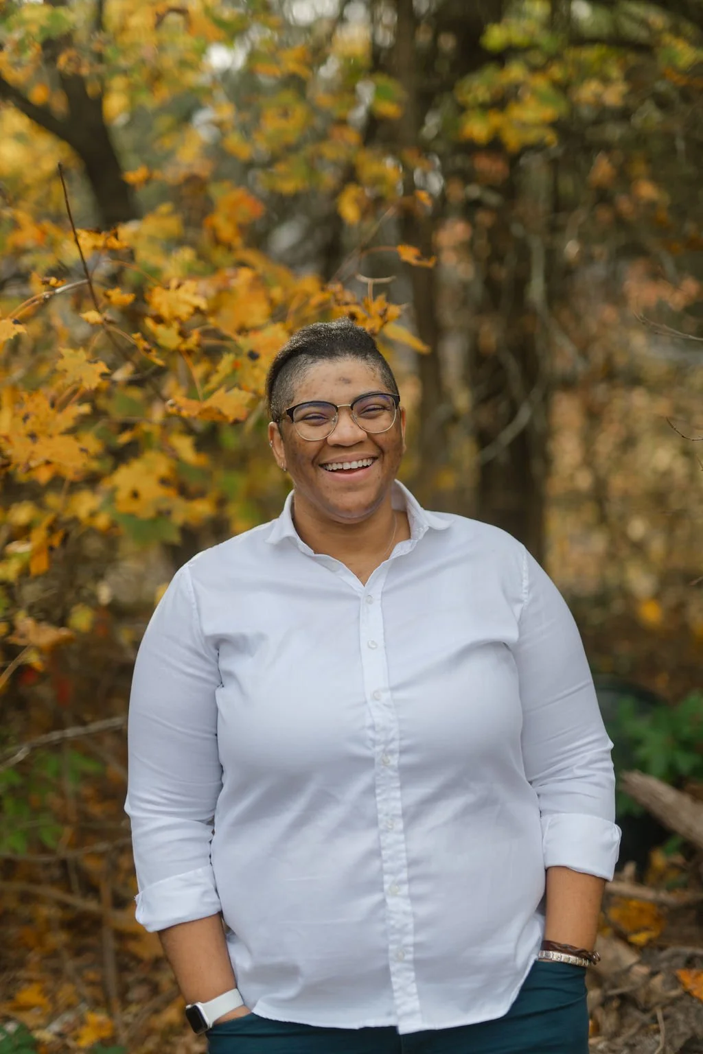 A smiling woman wearing glasses and a white button-up shirt standing outdoors in an autumnal setting with trees and yellow leaves in the background.