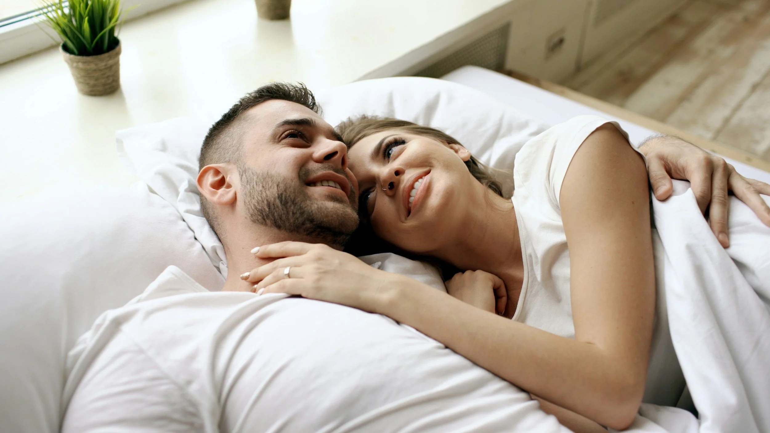 A couple lying in bed, smiling and looking at each other affectionately, with white bedding and a window in the background.