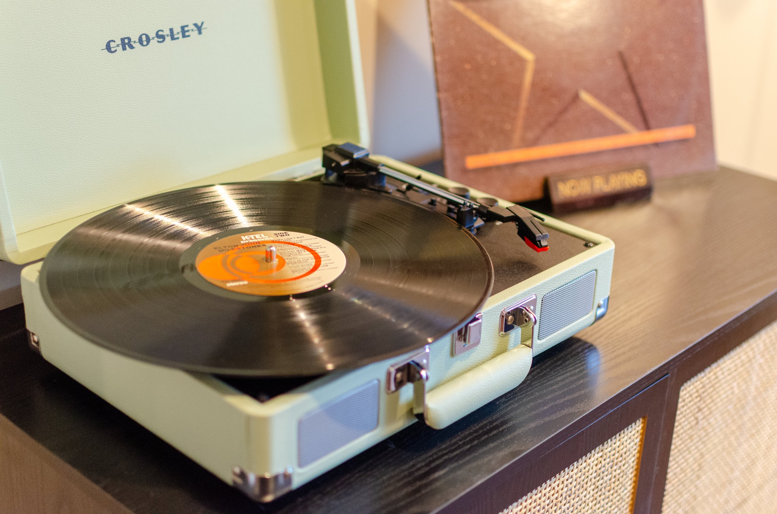 A vintage portable turntable playing a vinyl record on a dark wooden surface, with a light green case and a brown textured cover in the background.