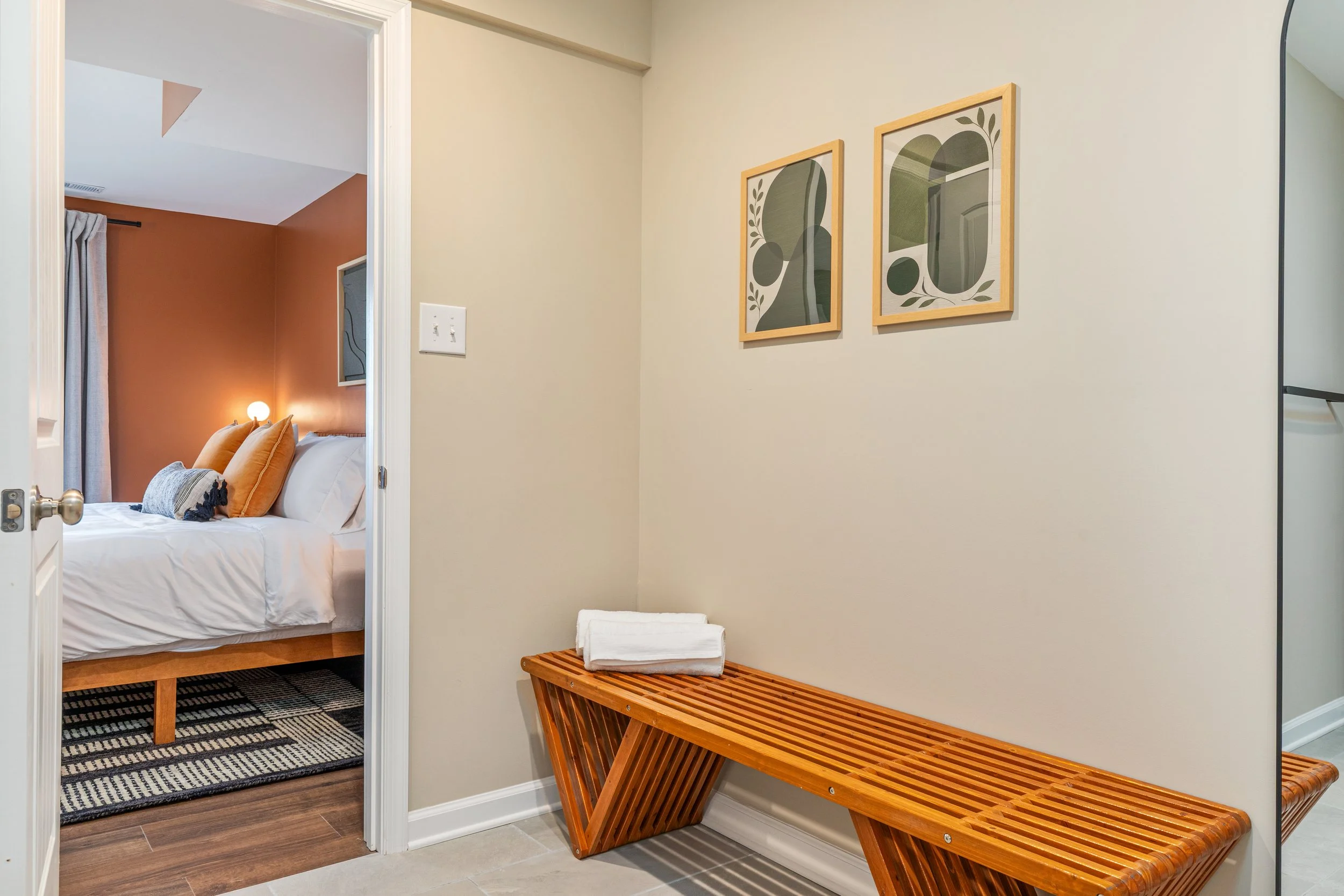 Interior view of a hallway with a wooden bench and two framed abstract art pieces on the wall, leading to a bedroom with a bed with white linens, orange pillows, and a bedside lamp.