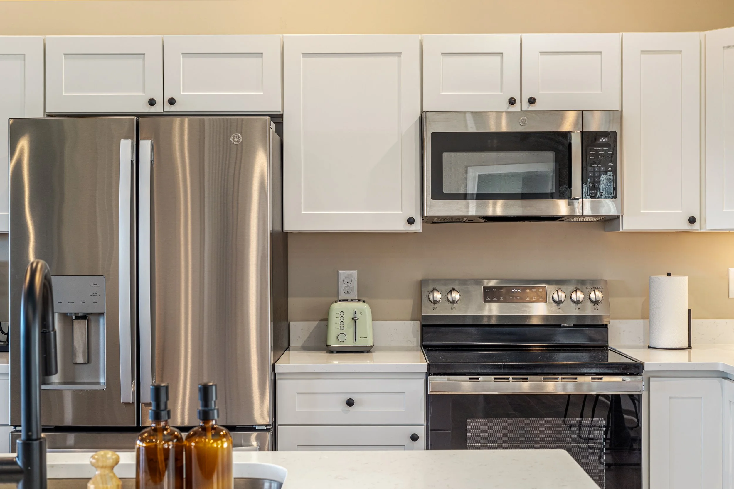 Modern kitchen with stainless steel appliances, white cabinets, a microwave, toaster, paper towel roll, and amber glass bottles on the counter.