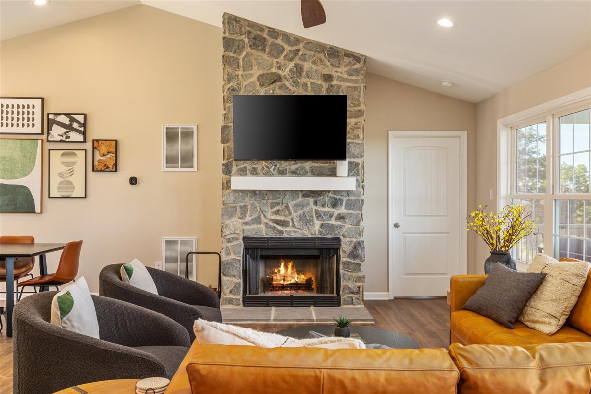 Living room with a stone fireplace, flat-screen TV above, yellow sofa, gray armchairs, and large windows with a vase of yellow flowers.