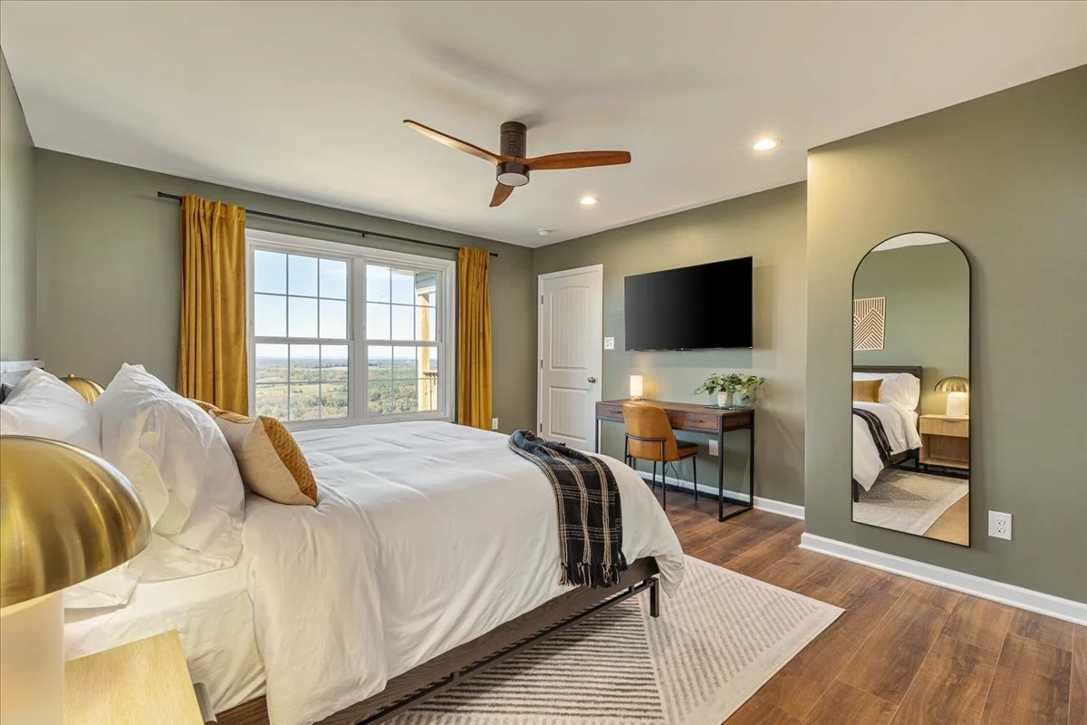 A bedroom with a large bed, gold lamps, a wooden desk with a chair, a wall-mounted TV, and a tall mirror reflecting part of the room. There are yellow curtains and a window showing a scenic view.
