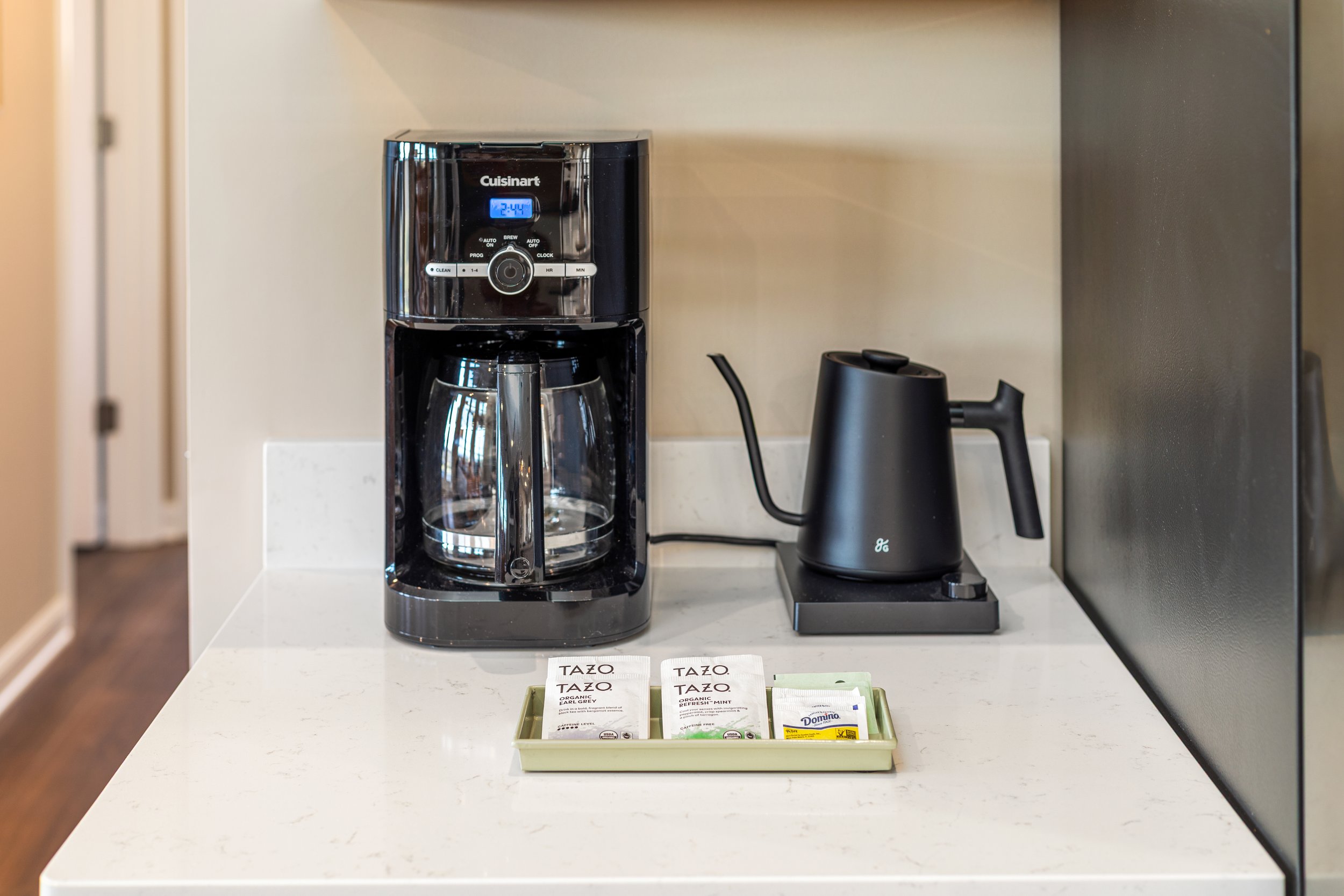 A black coffee maker and a black electric kettle on a white kitchen countertop, with a small tray of tea bags and sugar packets in front.