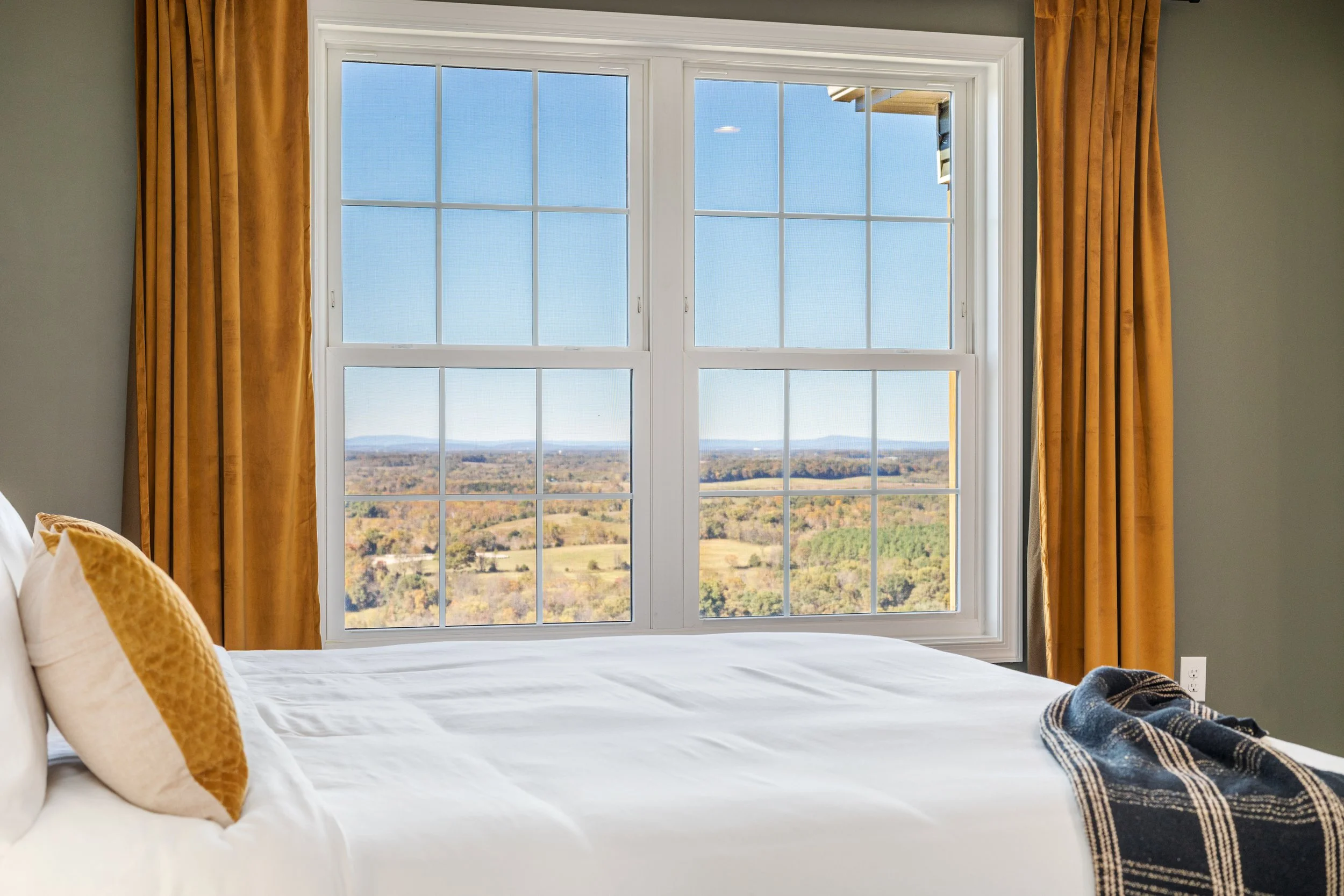 A bedroom with a large window showing a scenic landscape of fields and hills under a blue sky, flanked by mustard-yellow curtains, a bed with white bedding, and a plaid throw blanket.