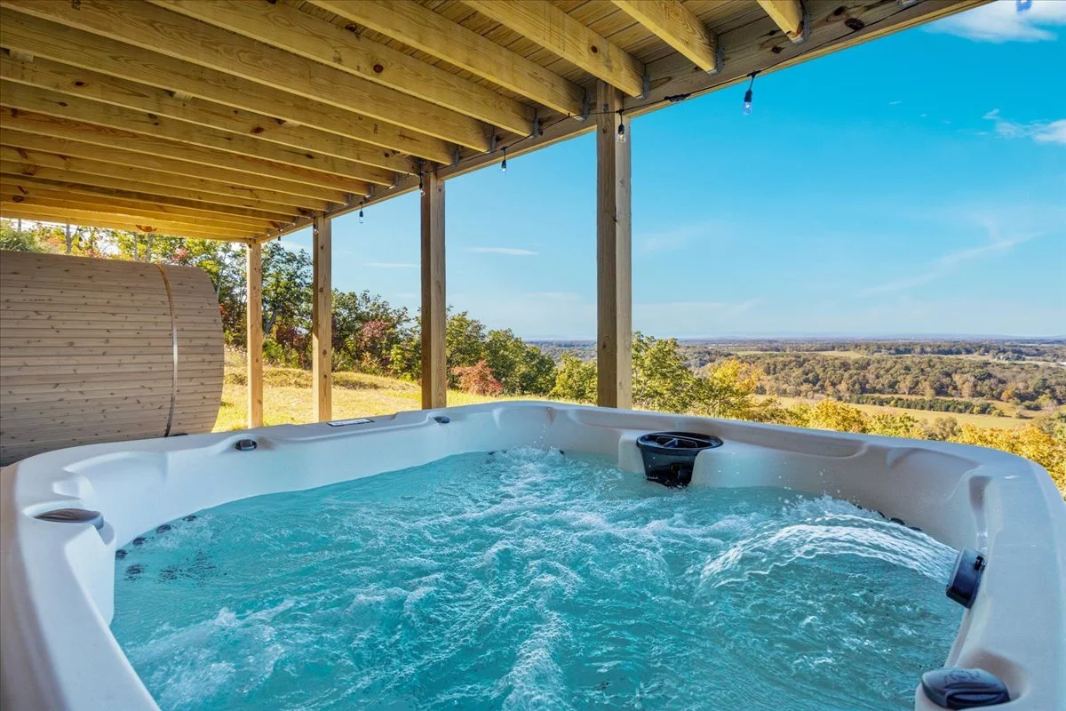 Outdoor hot tub with bubbling water on a wooden deck, overlooking a scenic view of trees and distant landscape under a clear blue sky.