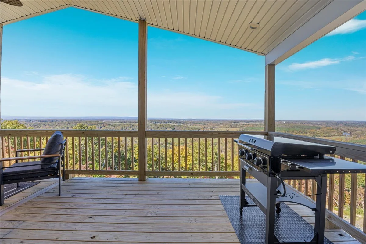 A balcony with a wooden floor, overlooking a scenic landscape of trees and a blue sky, furnished with a chair and a grill.