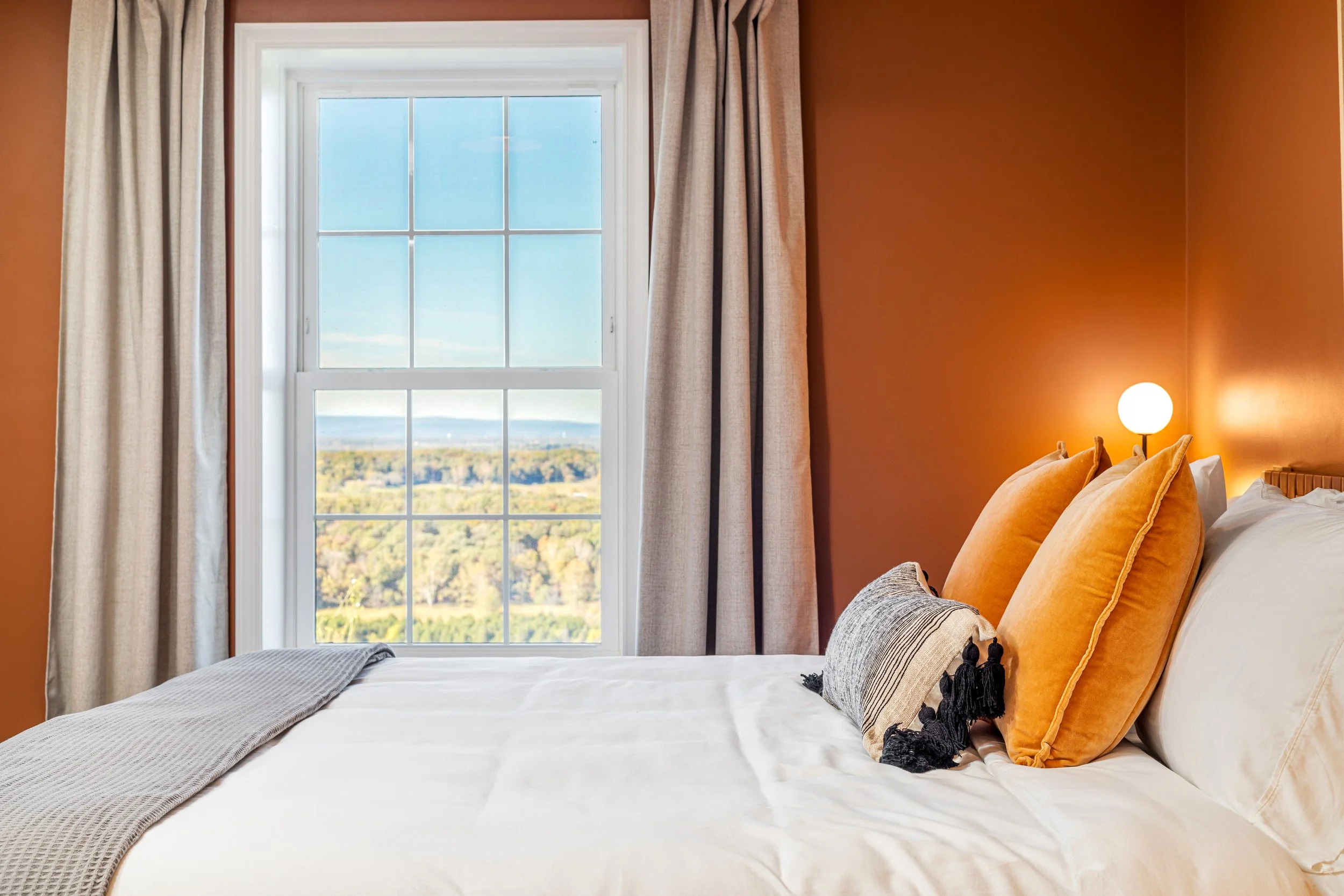 A bedroom with a large window showing a landscape view, with beige curtains, a bed with white sheets, two large mustard-colored pillows, a decorative pillow, and a small round wall sconce illuminating the wall.