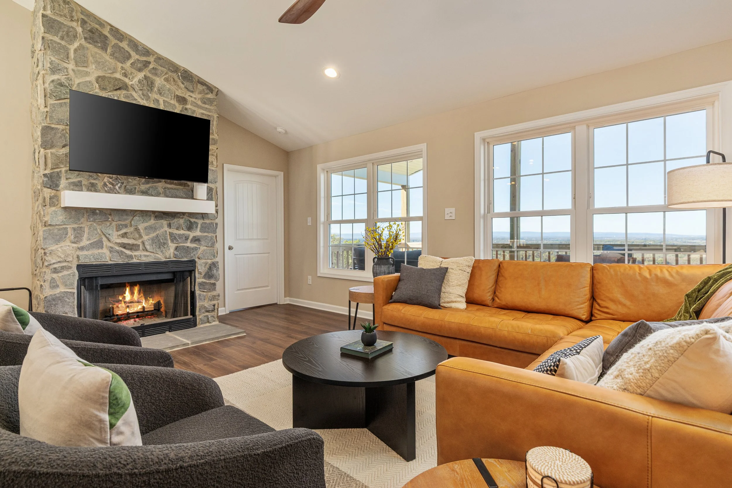 Living room with a fireplace and a mounted flat-screen TV on a stone wall. The room has a tan sofa, black armchairs, and large windows showing a scenic outdoor view. There's a black coffee table with a small plant, and a side table with a bouquet of yellow flowers. The room has wood flooring and beige walls.