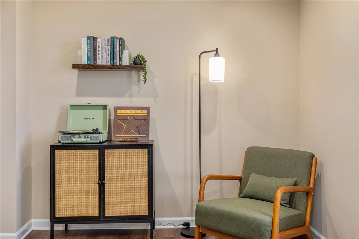 A cozy living room corner with a wooden armchair upholstered in green fabric and matching cushion. A black cabinet with rattan doors holds a vintage Crosley record player, an album cover, and a small decorative plant on top. Above, a wooden shelf displays a row of books and additional small plant. A tall white floor lamp provides warm lighting to the space.