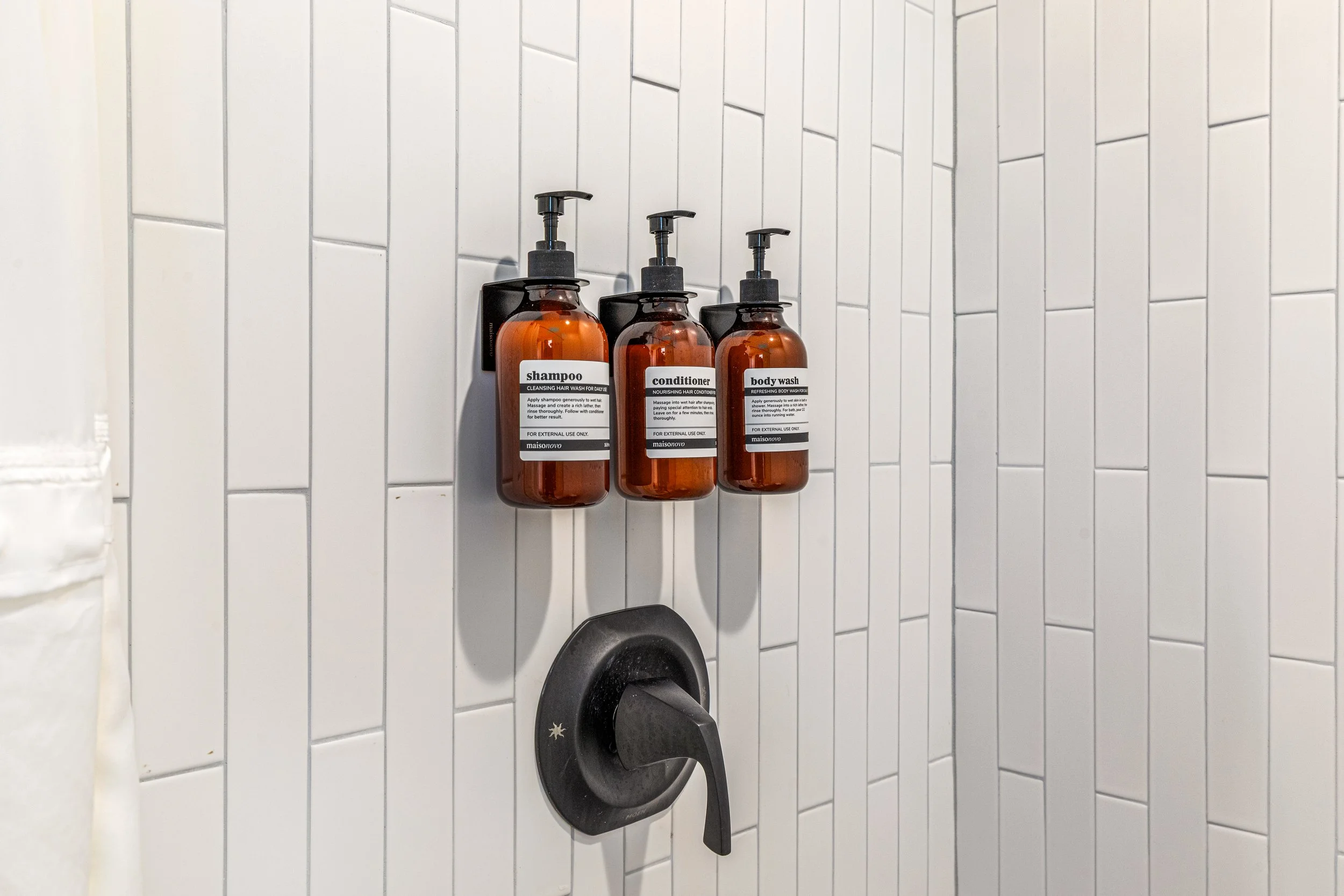 Three amber bottles with pump dispensers labeled shampoo, conditioner, and body wash mounted on a tiled shower wall.
