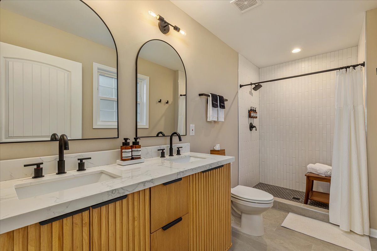 A modern bathroom with a double vanity featuring a white marble top and black fixtures, a large mirror with arched edges, a separate shower area with white tiles, a robe hook, a small wooden bench, and a window with white trim.