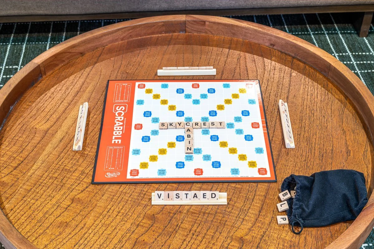Scrabble game on a round wooden table with tiles spelling S. K. Y. C. R. E. S. T., B. I. P., and V. I. S. T. A. E. D., a cloth bag with more tiles, and a tiled game board with blue, yellow, red, and white squares.