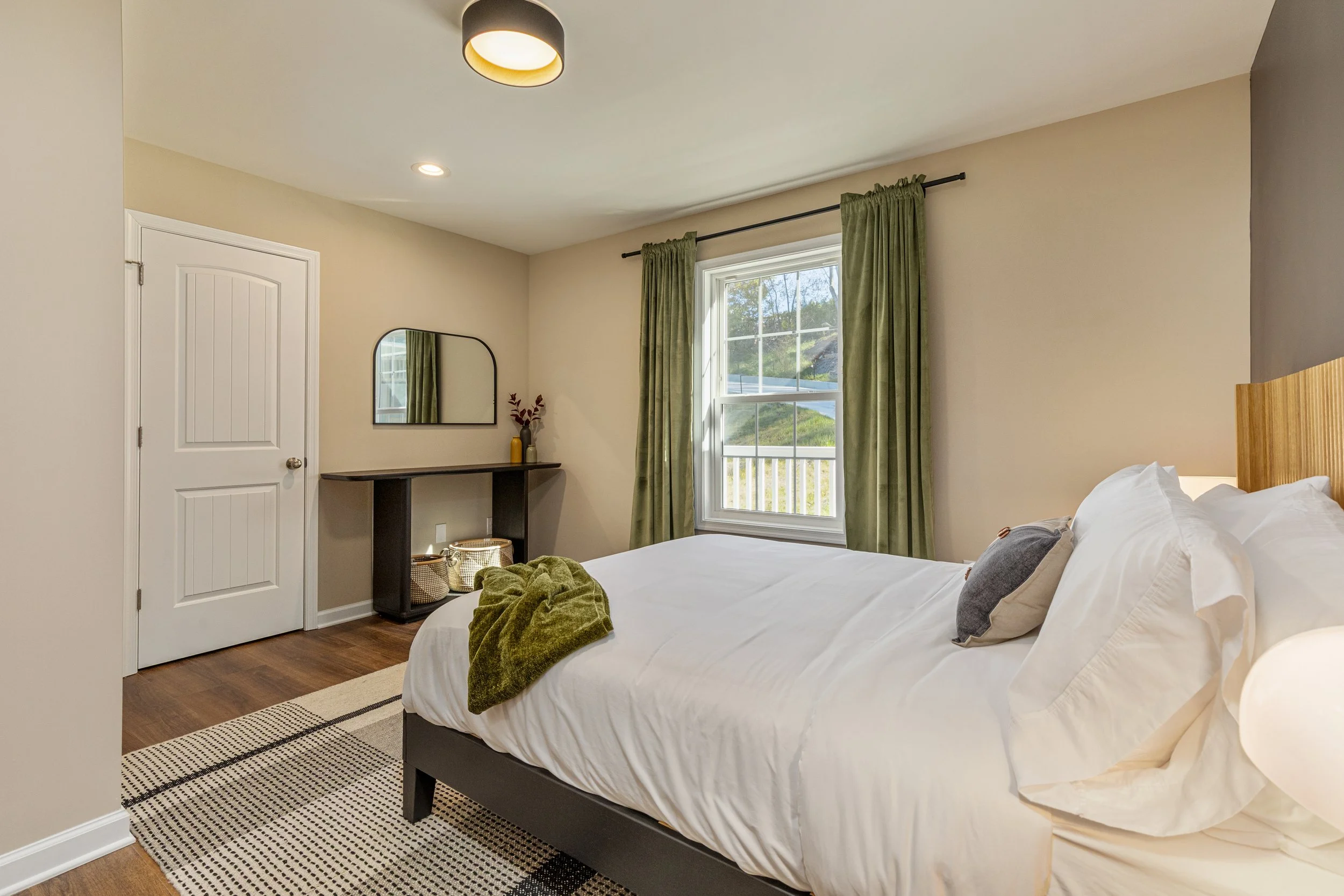 A bedroom with a bed, green curtains, a window showing an outdoor view, a mirror, a dresser with a vase, and a lamp.