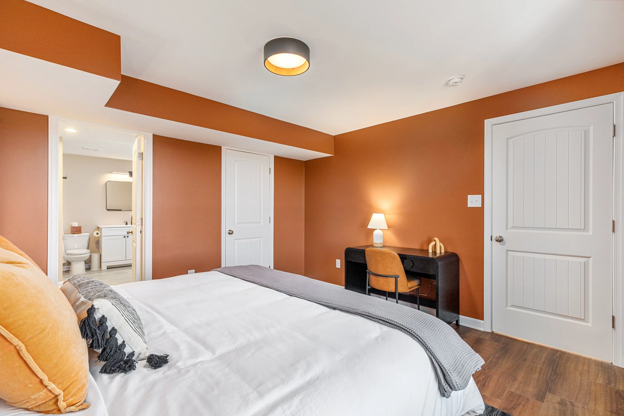 A bedroom with a bed, a desk with a lamp, and two closed white doors, with an attached bathroom visible through an open door.