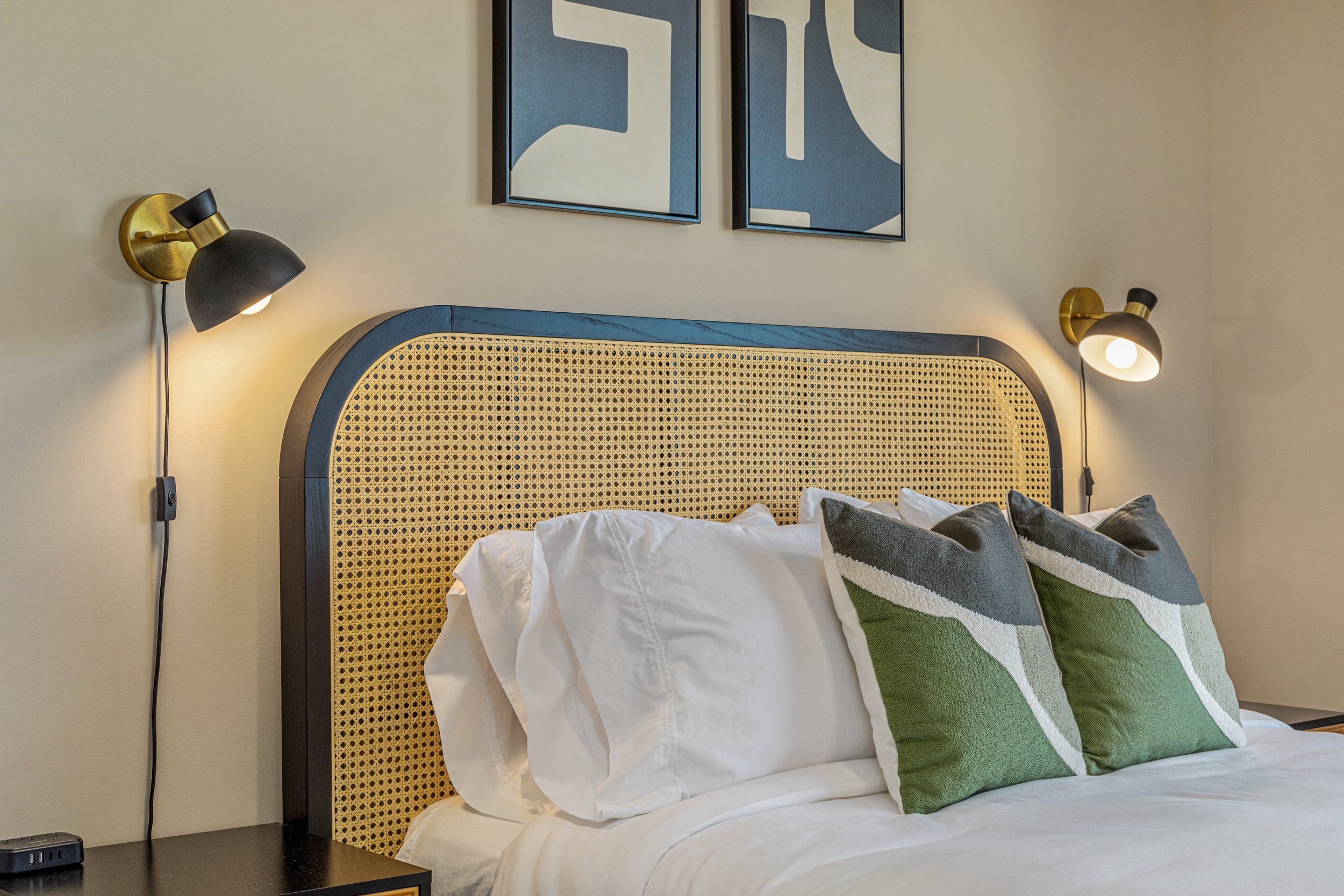 A bedroom with a bed featuring a rattan headboard, white bedding, and green and gray pillows. Two wall-mounted lamps are on either side of the bed, and framed abstract art hangs above.