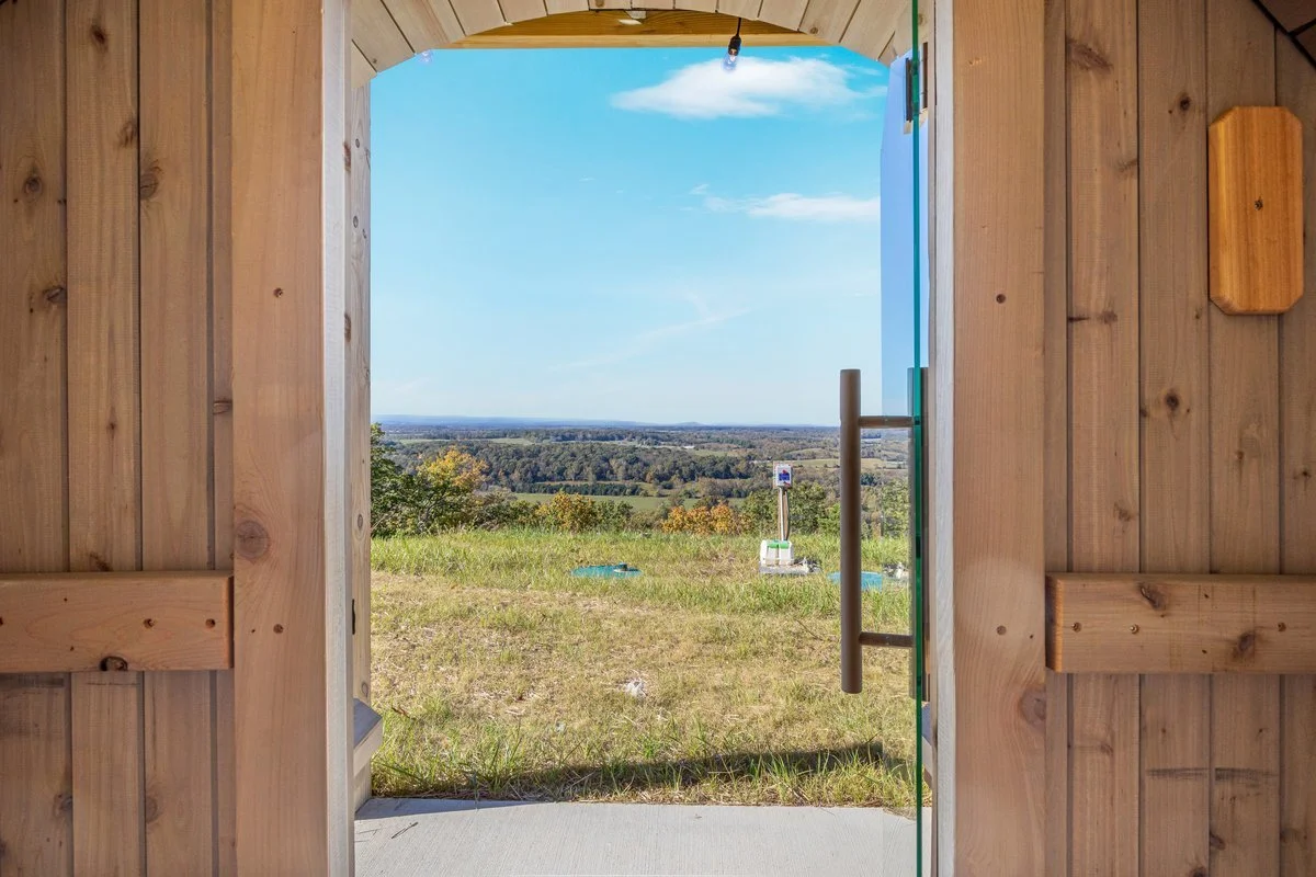 View through a wooden door revealing a landscape of green fields and trees under a blue sky with scattered clouds.
