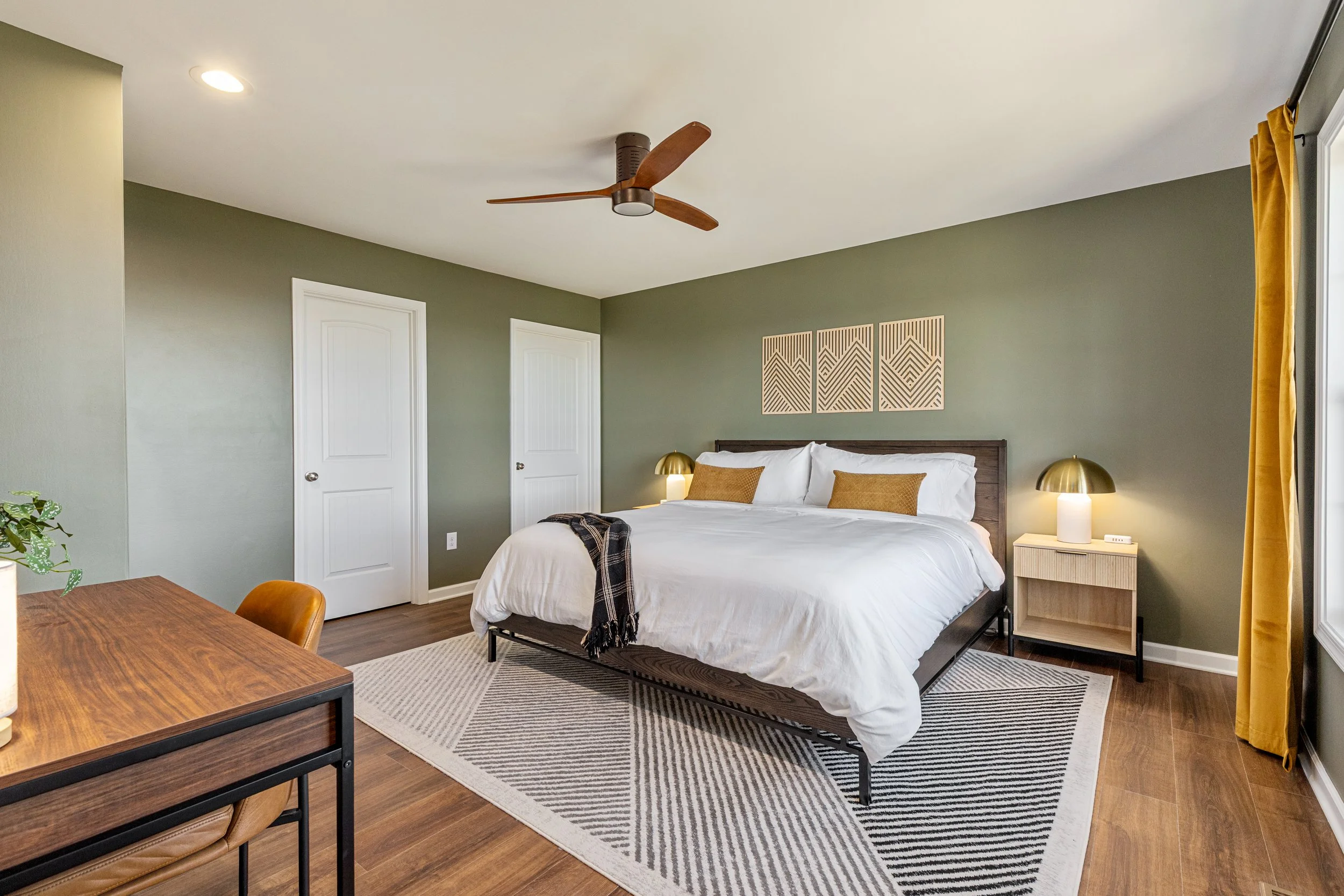 A bedroom with a green wall, white bed, yellow pillows, and wooden nightstands with lamps. A black and white rug is beneath the bed, and a wooden ceiling fan hangs from the ceiling.