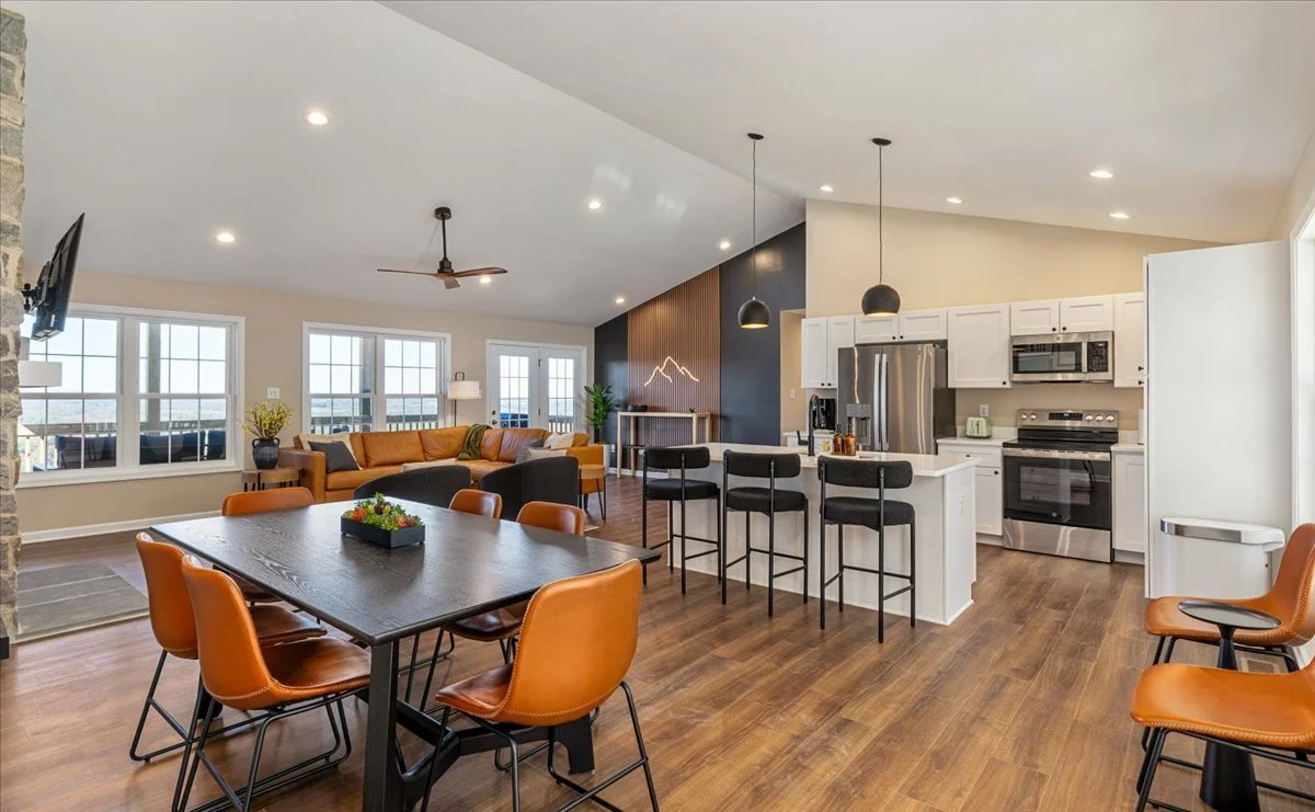 Open-concept living room, dining area, and kitchen with hardwood floors, white cabinetry, stainless steel appliances, black barstools, and large windows with natural light.