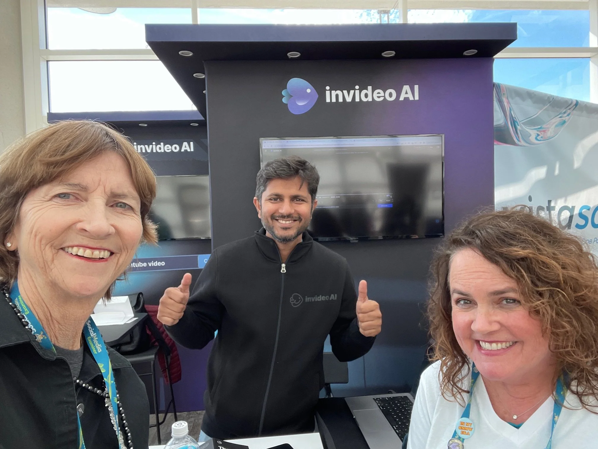 Three smiling people, two women and one man, posing for a photo at an event booth with a large screen and a sign that reads 'invideo AI' behind them.