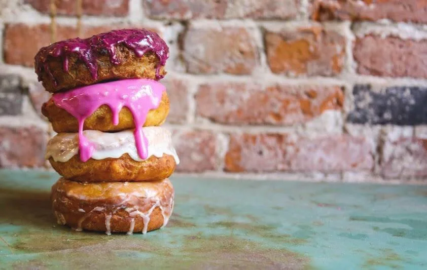 Stack of four donuts with pink, white, and purple icing, set against a rustic brick wall background. Donuts to Ayurveda.