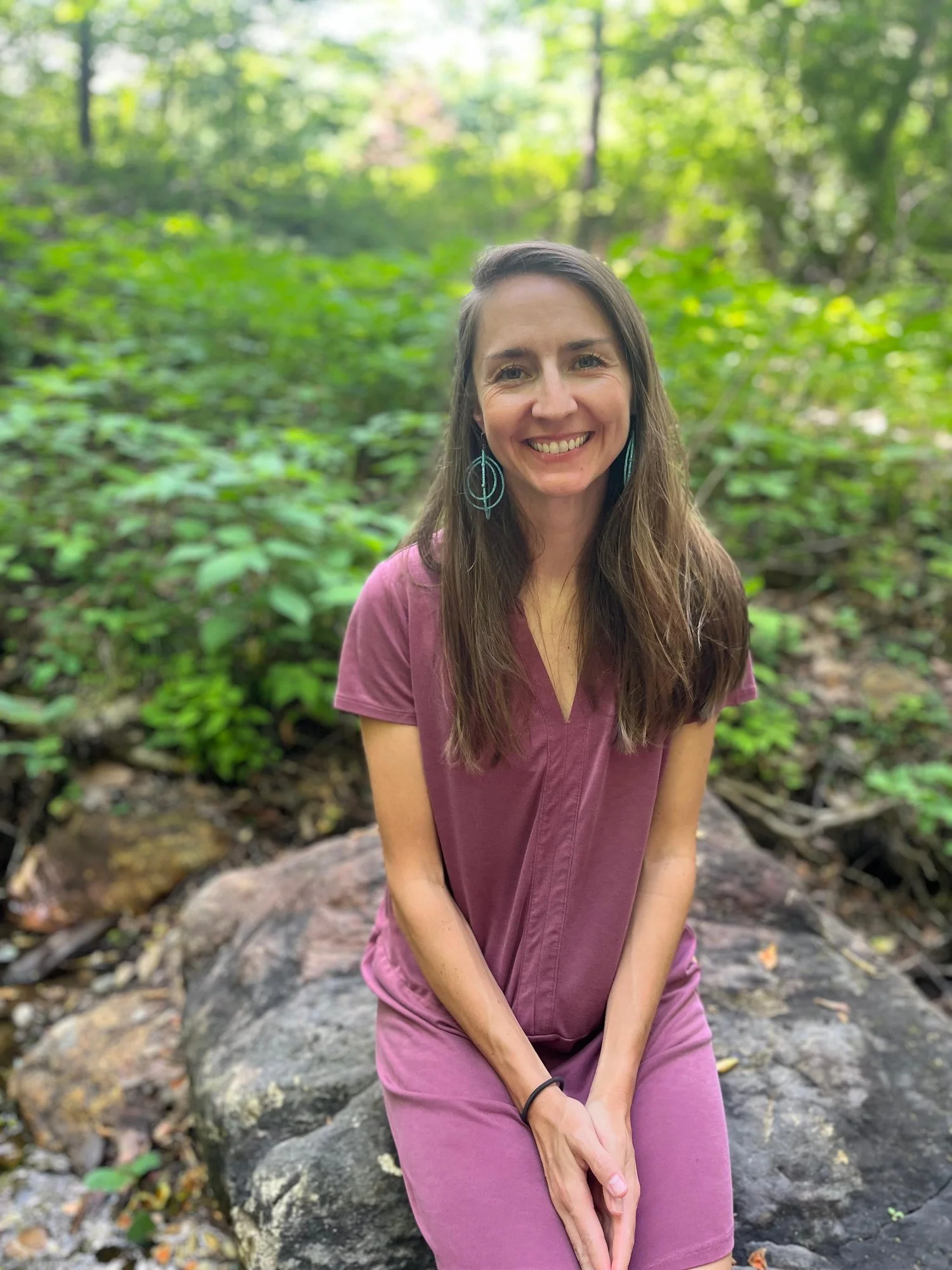 A woman with long brown hair smiling while sitting on a rock in a green forest. Alex Rice, Ayurvedic Practitioner.