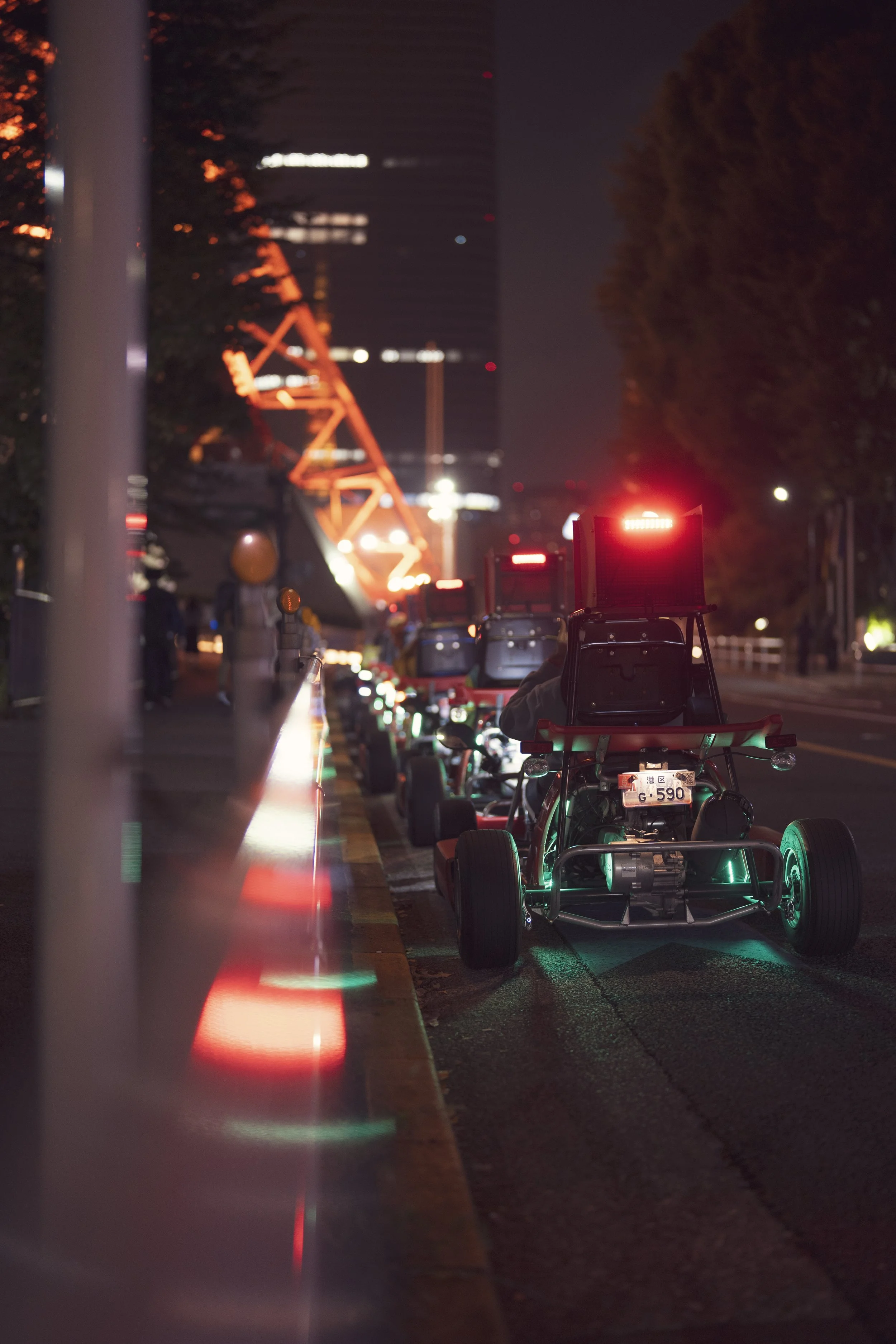 Row of go-karts with illuminated lights parked along a city street at night, with a ferris wheel and tall buildings in the background.
