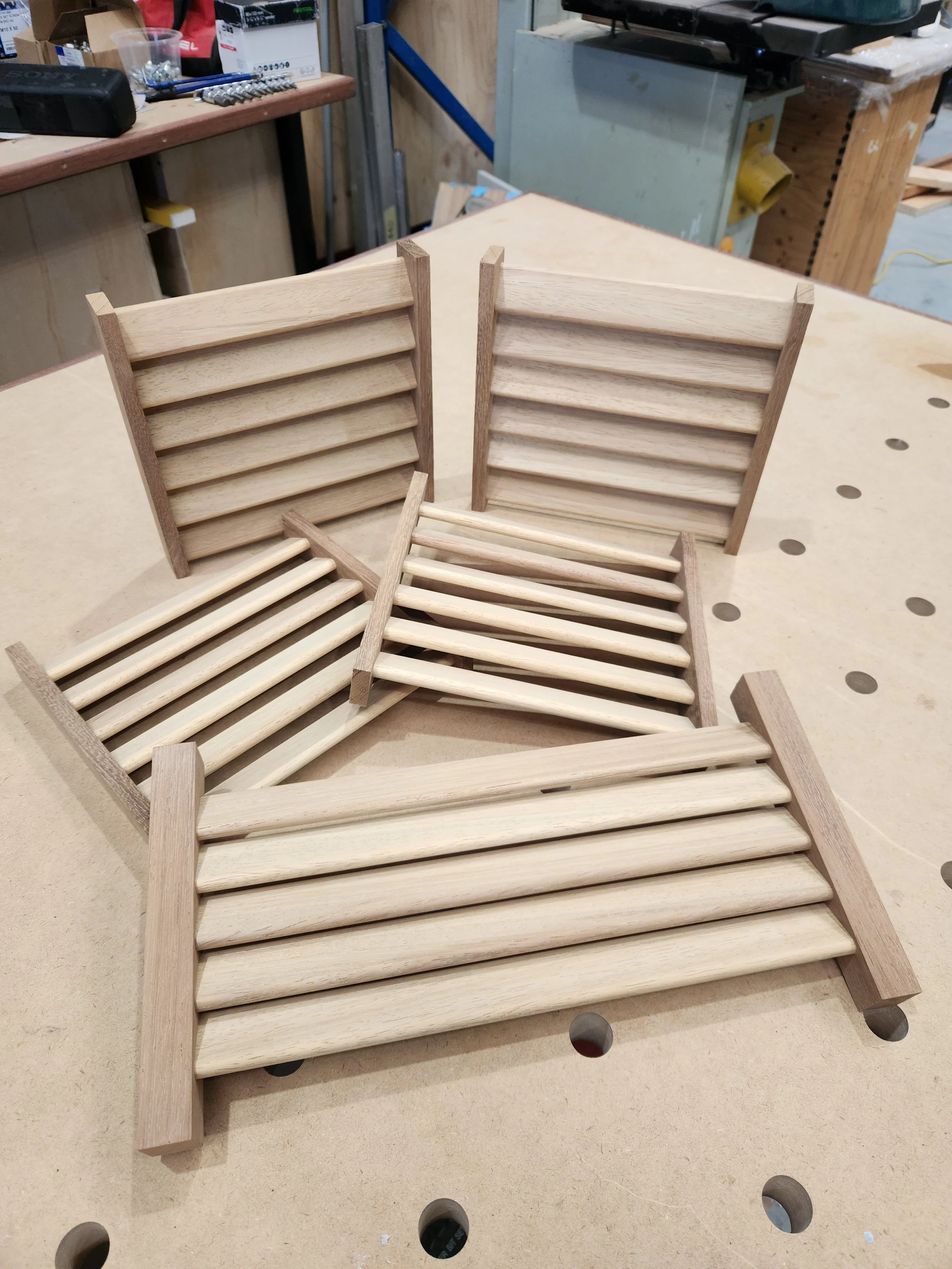 Three unfinished wooden slatted benches or shelves in a woodworking shop, with tools and equipment in the background.