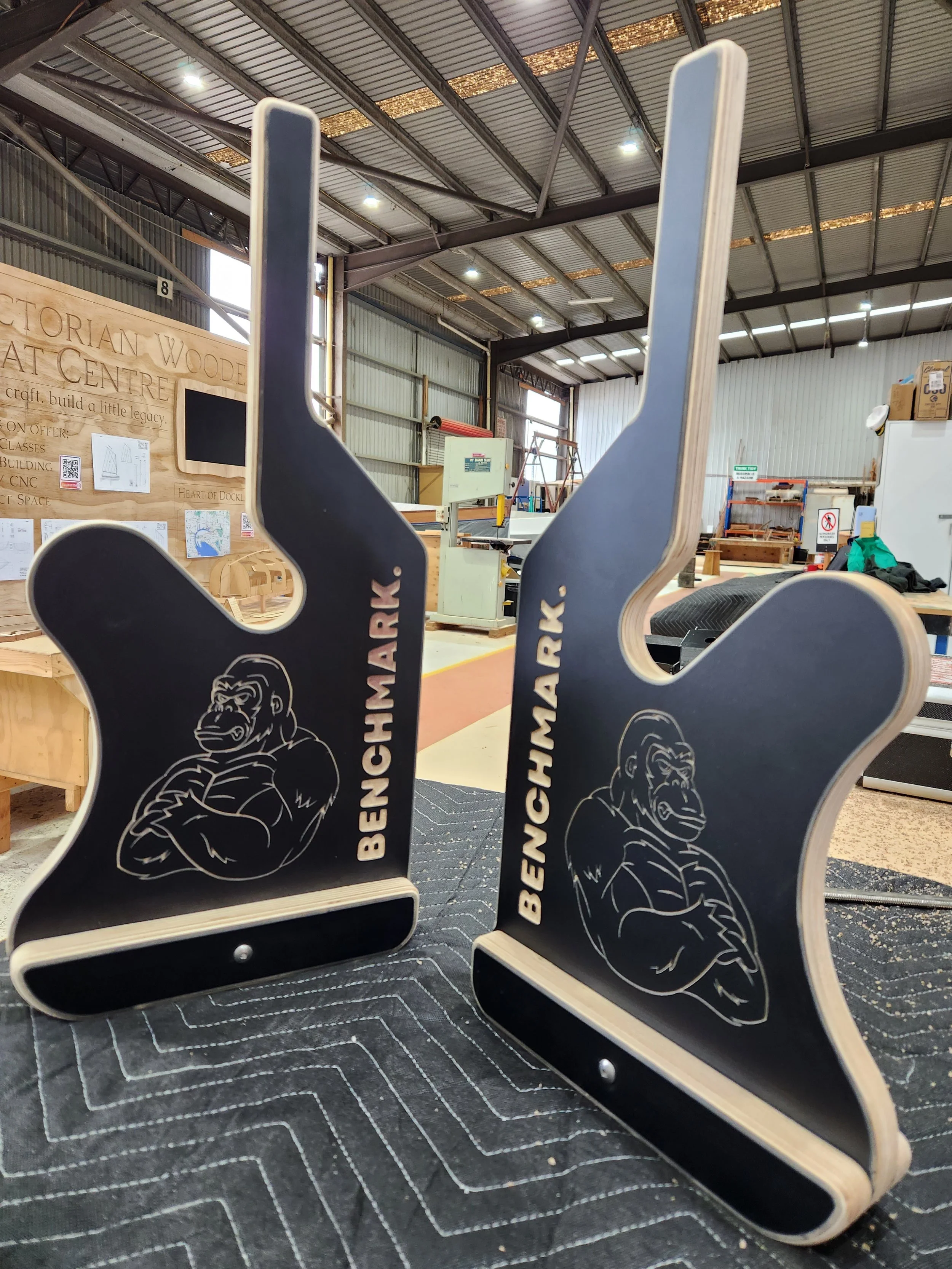 A close-up image of black wood and metal tongs with the word "BENCHMARK" and a gorilla logo printed on them, placed on a circular black base, in a workshop or warehouse with industrial equipment and wood projects in the background.
