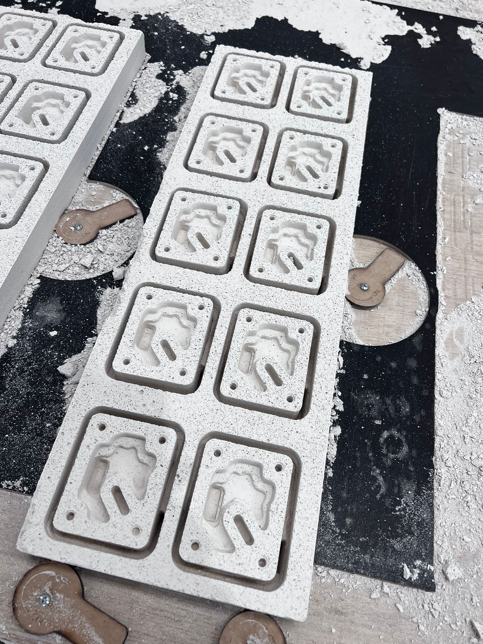 Several white concrete molds with indentations of a brick shape and a chain link attached to circular wooden disks, on a work surface with white powder and black surface.