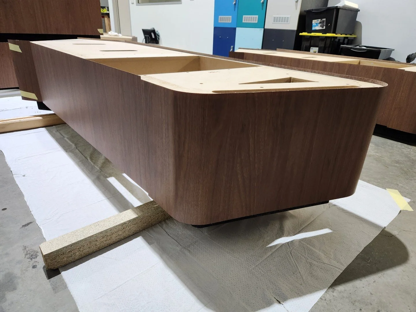 Large wooden desk with a dark wood finish, set on styrofoam blocks inside a workshop or storage area, with lockers and various equipment in the background.