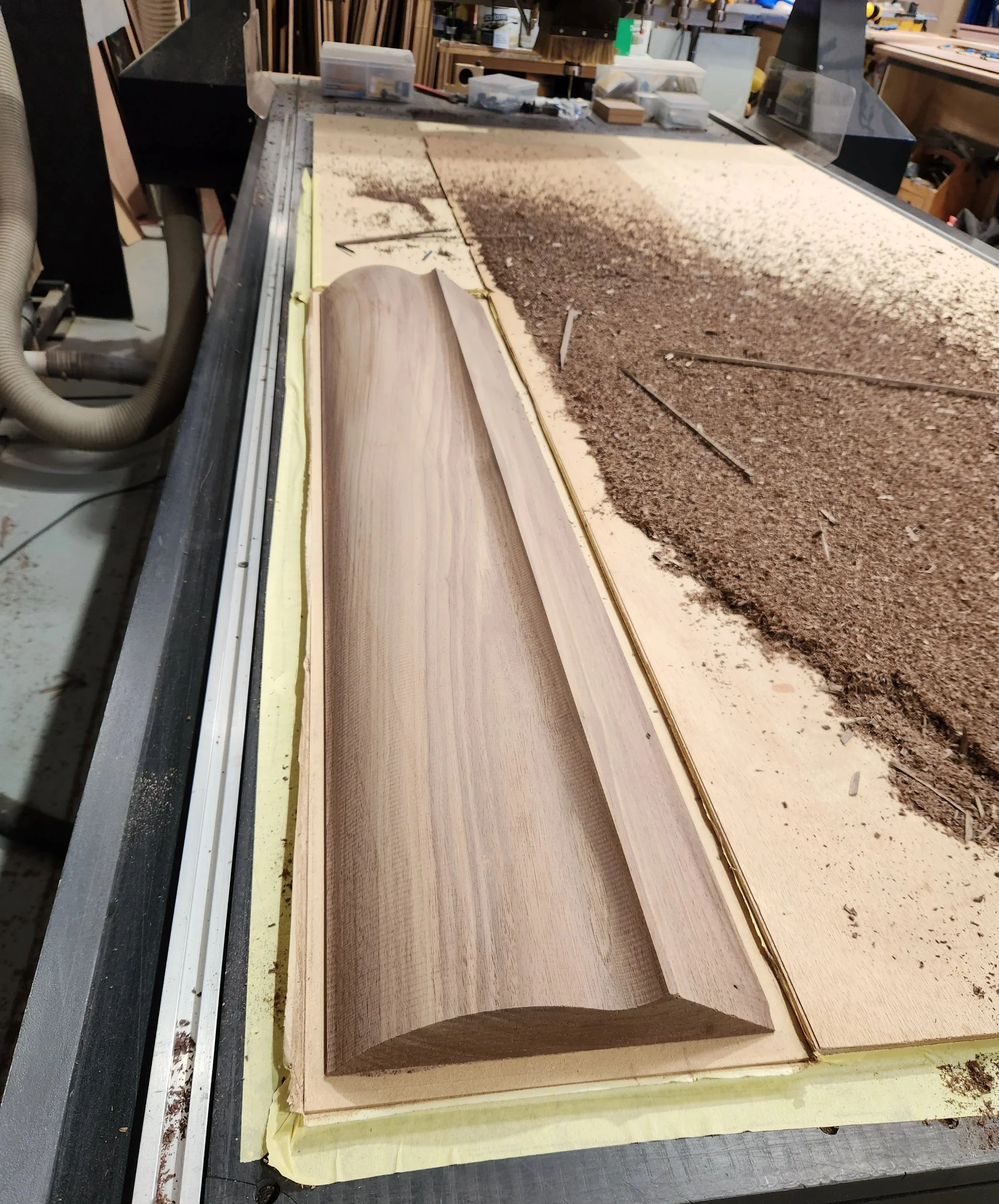 A woodworking workbench in a carpentry shop with a piece of wood being shaped, surrounded by sawdust, tools, and supplies.