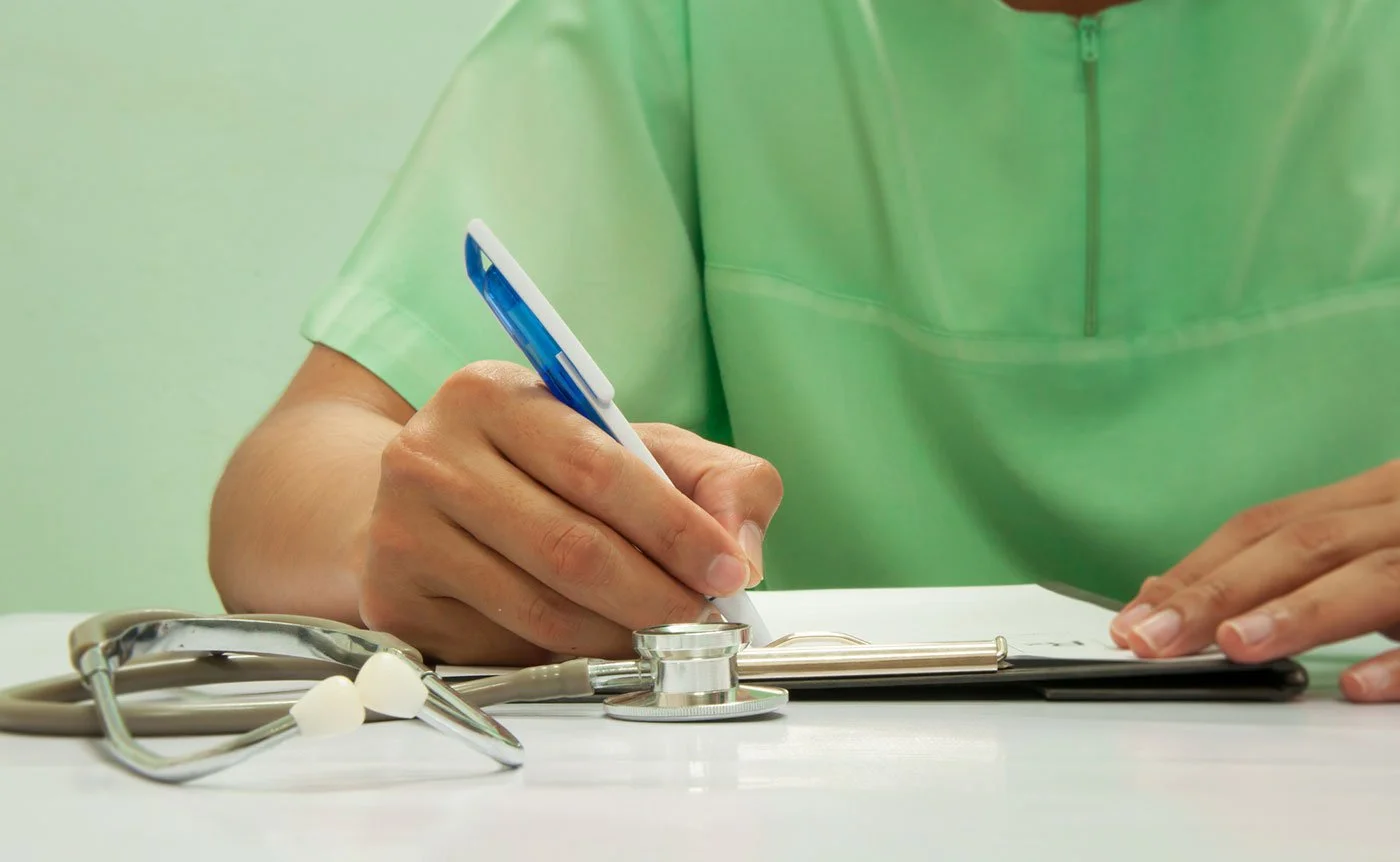 Close-up of a healthcare professional wearing a green scrub, writing on a clipboard, with a stethoscope placed on the white surface beside them.