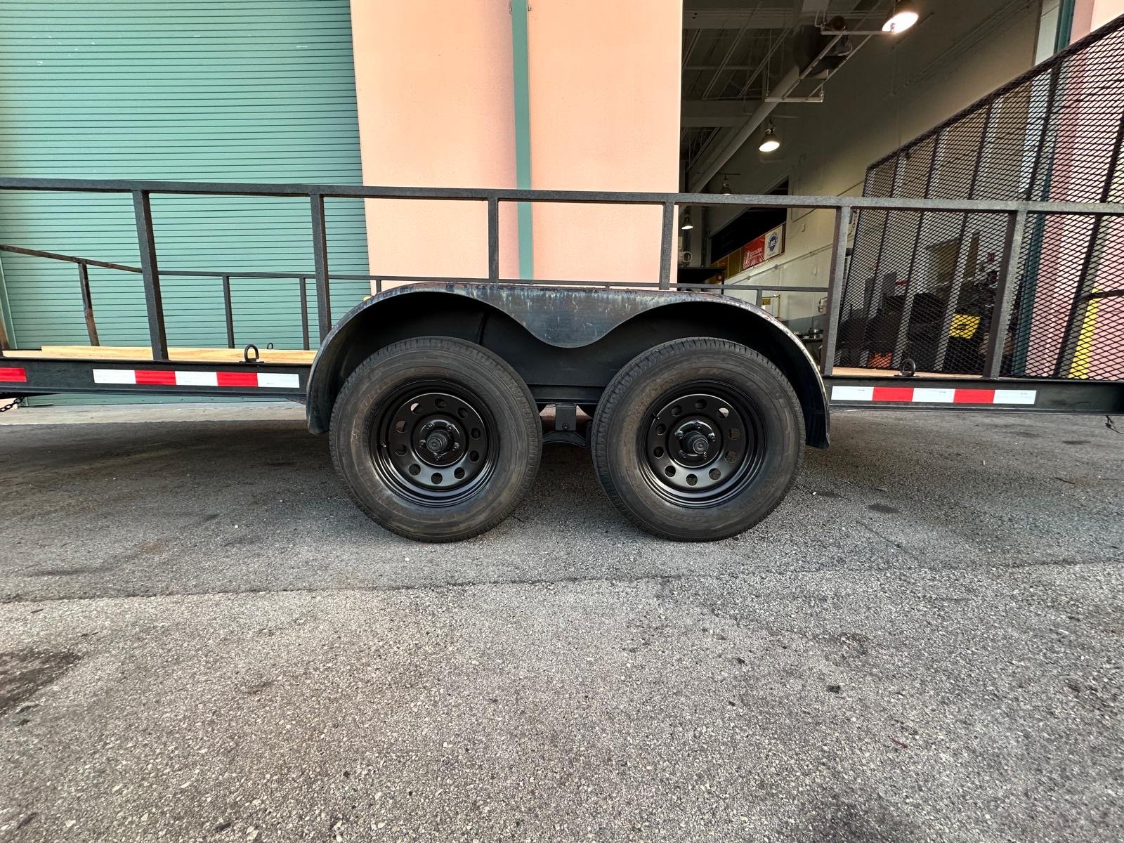 A flatbed trailer with two black tires and metal side rails, parked on a gravel surface in front of a building with teal and pink walls and an open garage area.