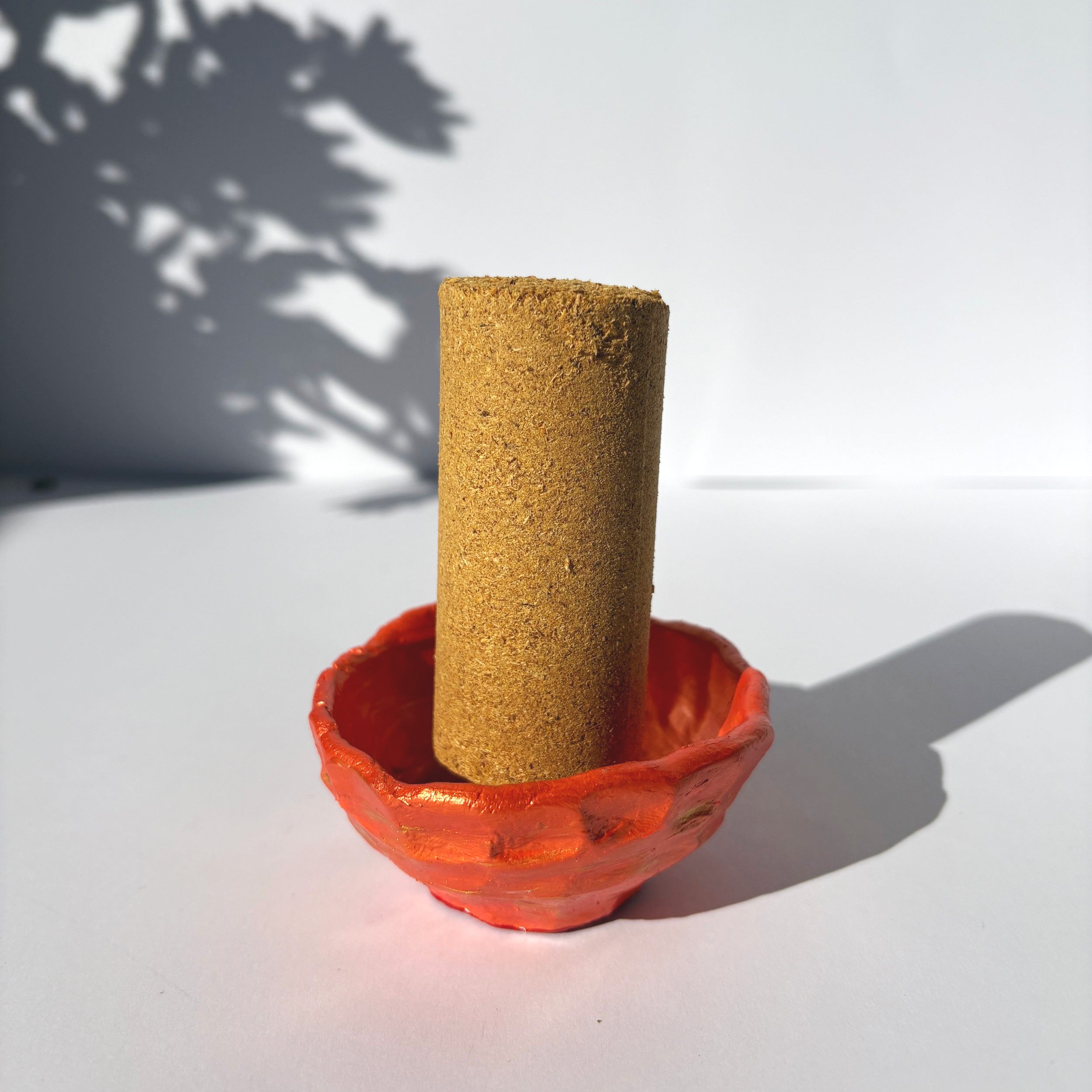 Handcrafted copper clay bowl holding a thick incense cone, with a soft shadow of leaves cast on the background.