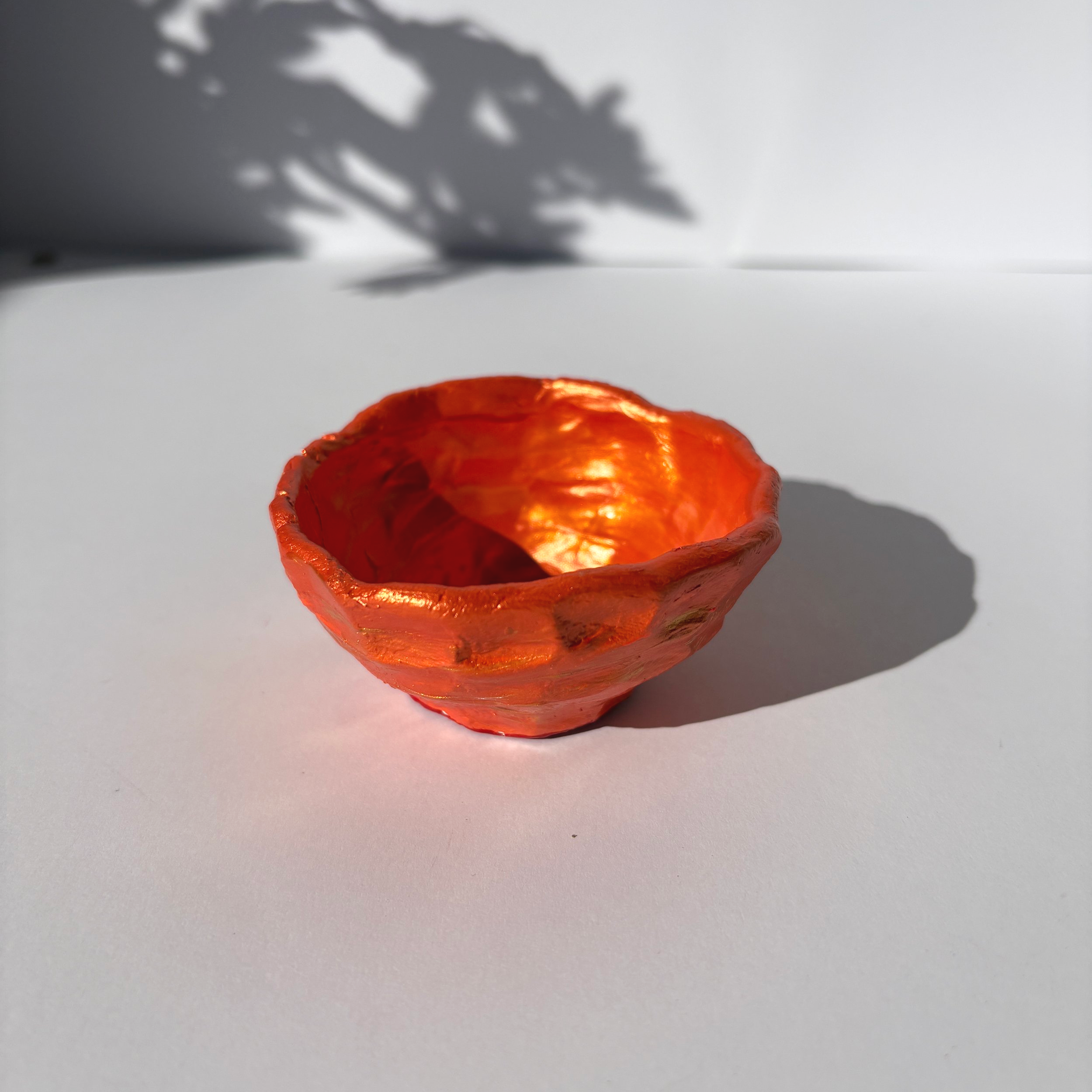 Empty handmade clay bowl painted in vibrant copper tones, displayed in sunlight with leafy shadows behind.