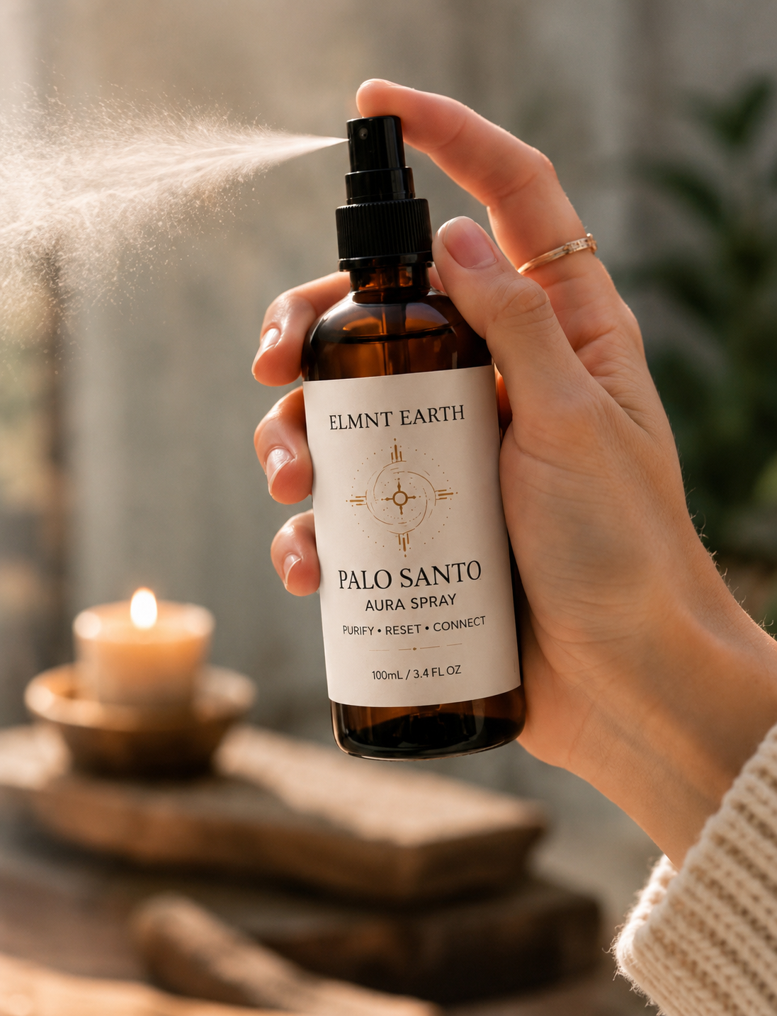 Person holding a brown spray bottle labeled 'Elmtn Earth Palo Santo Aura Spray,' spraying mist in a cozy setting with a lit candle and natural decor in the background.
