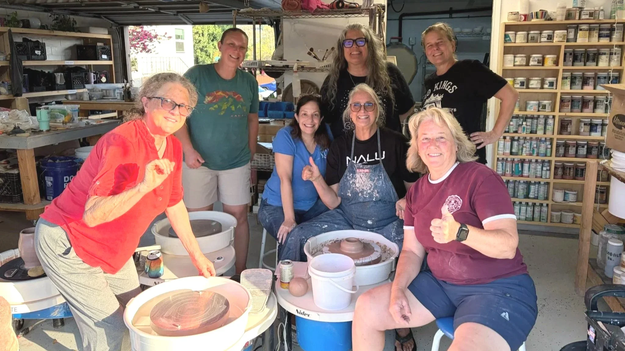 A group of eight women at SD Mad Mudders pottery studio in Spring Valley, California, some sitting and some standing, smiling and giving thumbs up. There are pottery wheels, shelves with paint bottles, and tools in the background.