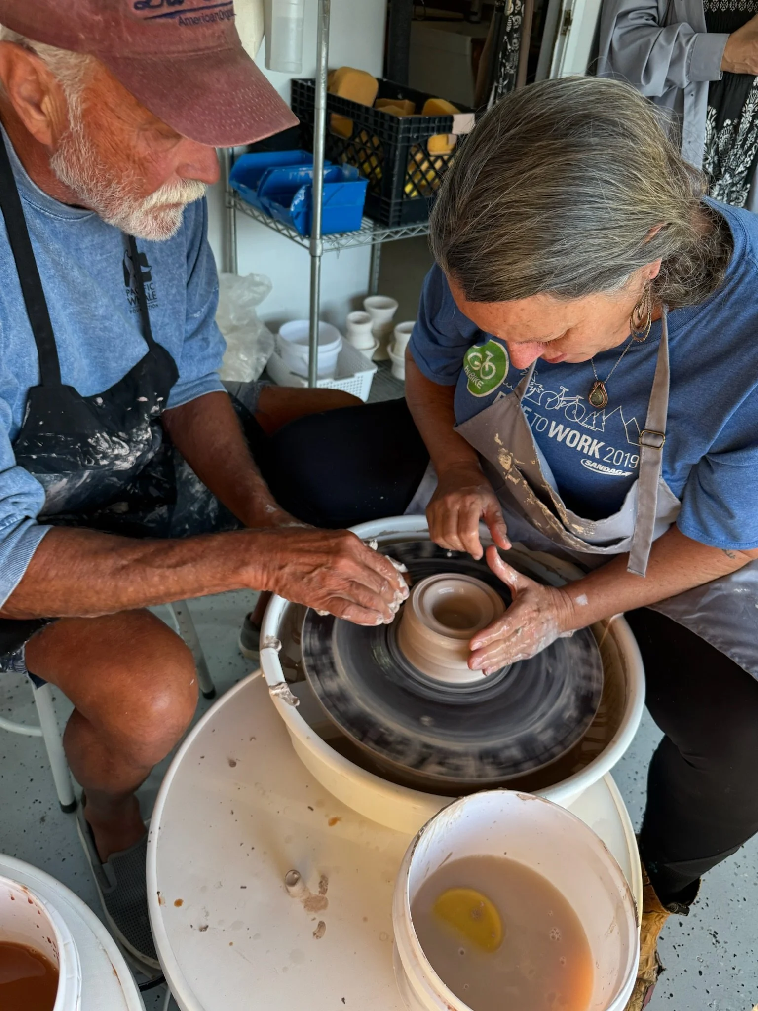 Mark Reed Heckl teaches a wheel throwing class working at SD Mad Mudders pottery studio in Spring Valley, California. He guides a student as she opens a small cylinder. There are pottery tools and buckets with clay and water nearby.