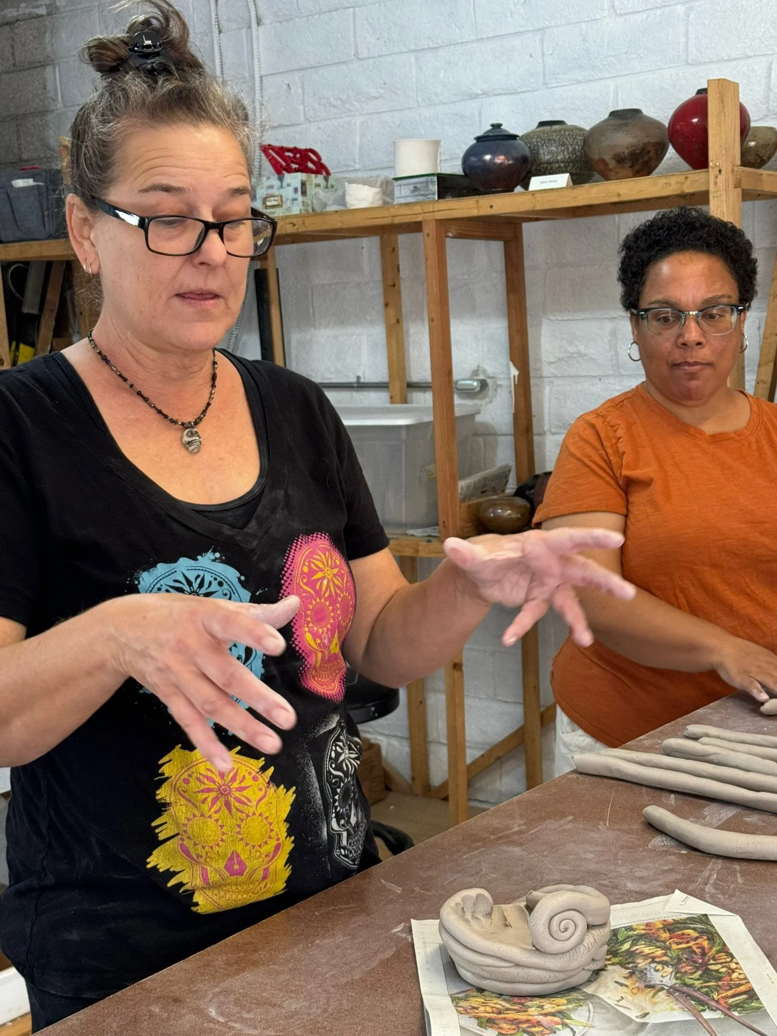 Sara DuVall teaches a hand-building class at SD Mad Mudders pottery studio in Spring Valley, California. A student watches as she explains coil techniques to a group class. In the background there are shelves holding studio member tools and pieces.