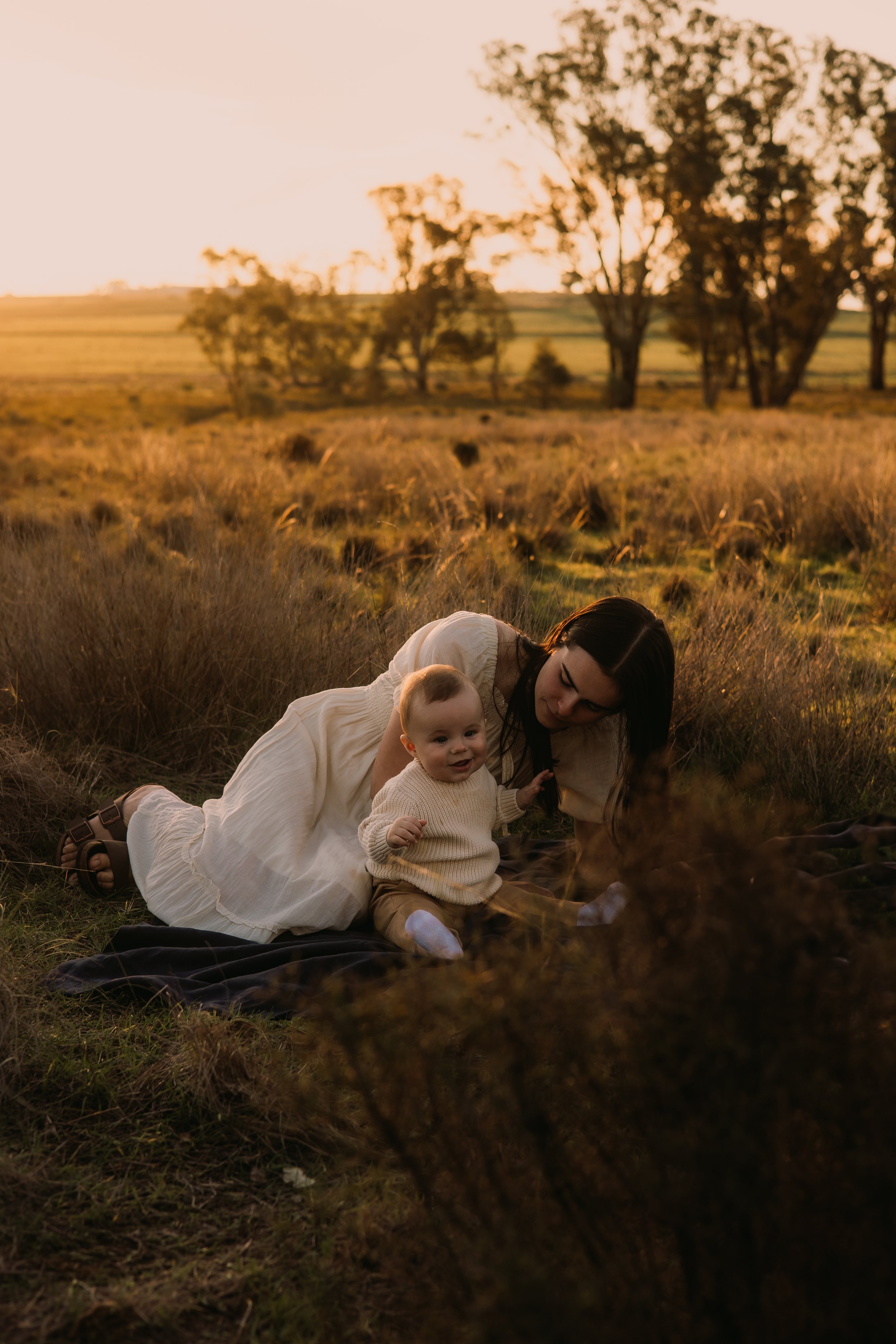 A woman and three children sitting on a blanket outdoors with vintage-style clothing, surrounded by flowers and baskets, under a blanket with lace details.