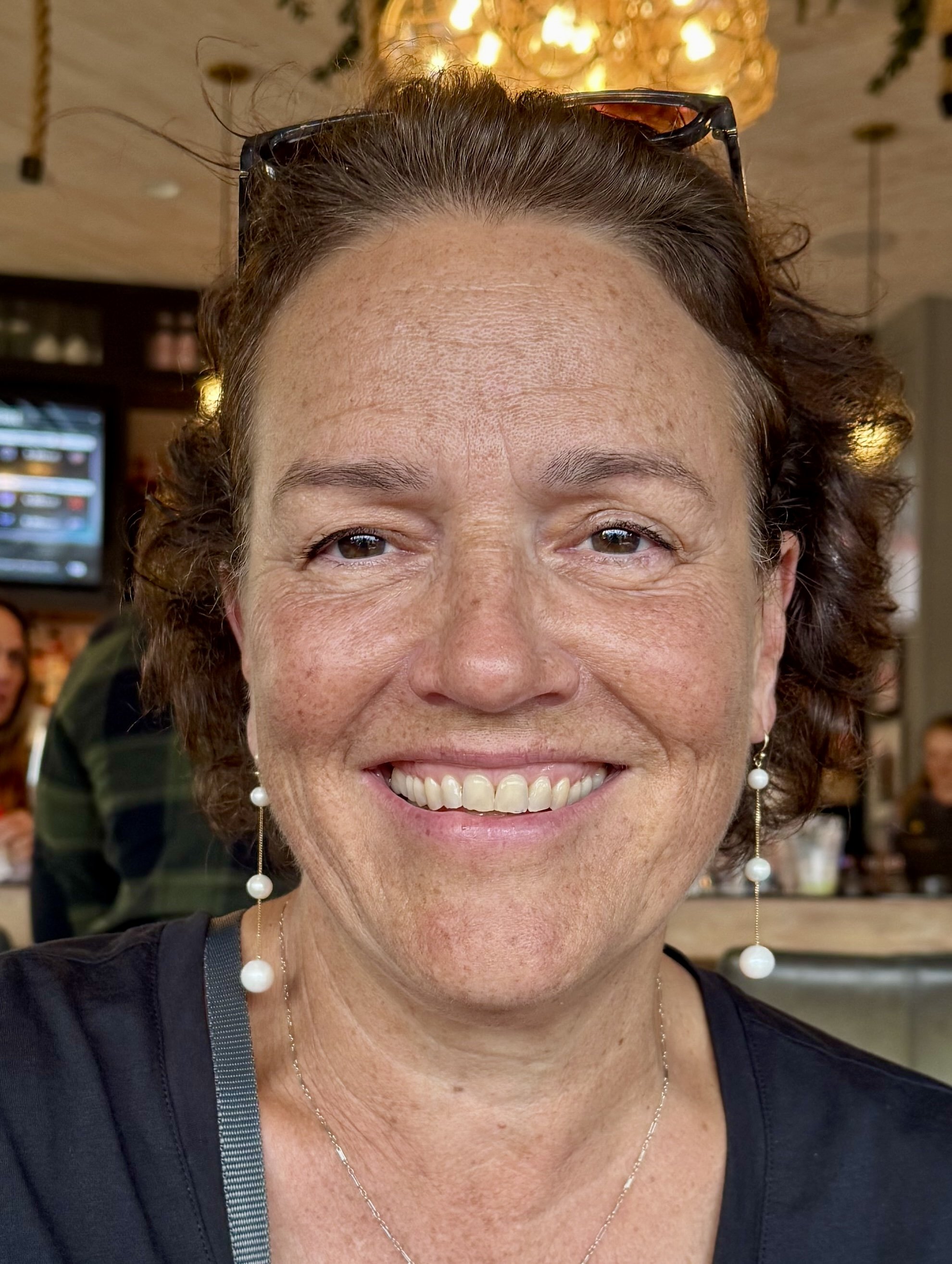 A smiling woman with short, curly brown hair and pearl earrings, wearing glasses on her head, in a restaurant or café with a blurry background of other people and a chandelier.