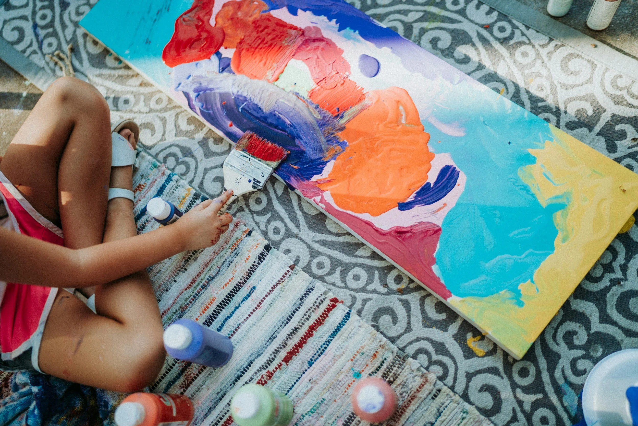 Child sitting on a striped rug painting an abstract picture on a large canvas with bright colors including orange, blue, yellow, and purple, surrounded by bottles of paint.