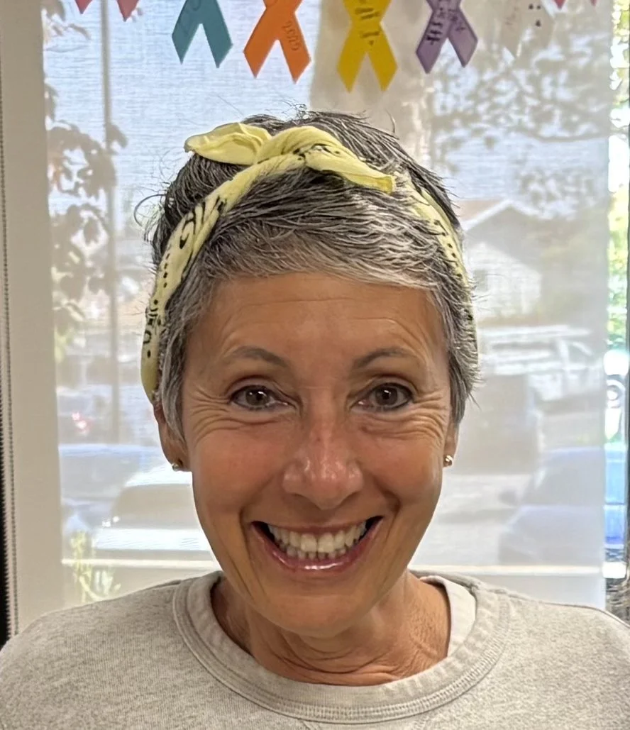 A smiling woman with short gray hair wearing a yellow headband, standing in front of a window with colorful ribbons hanging above.